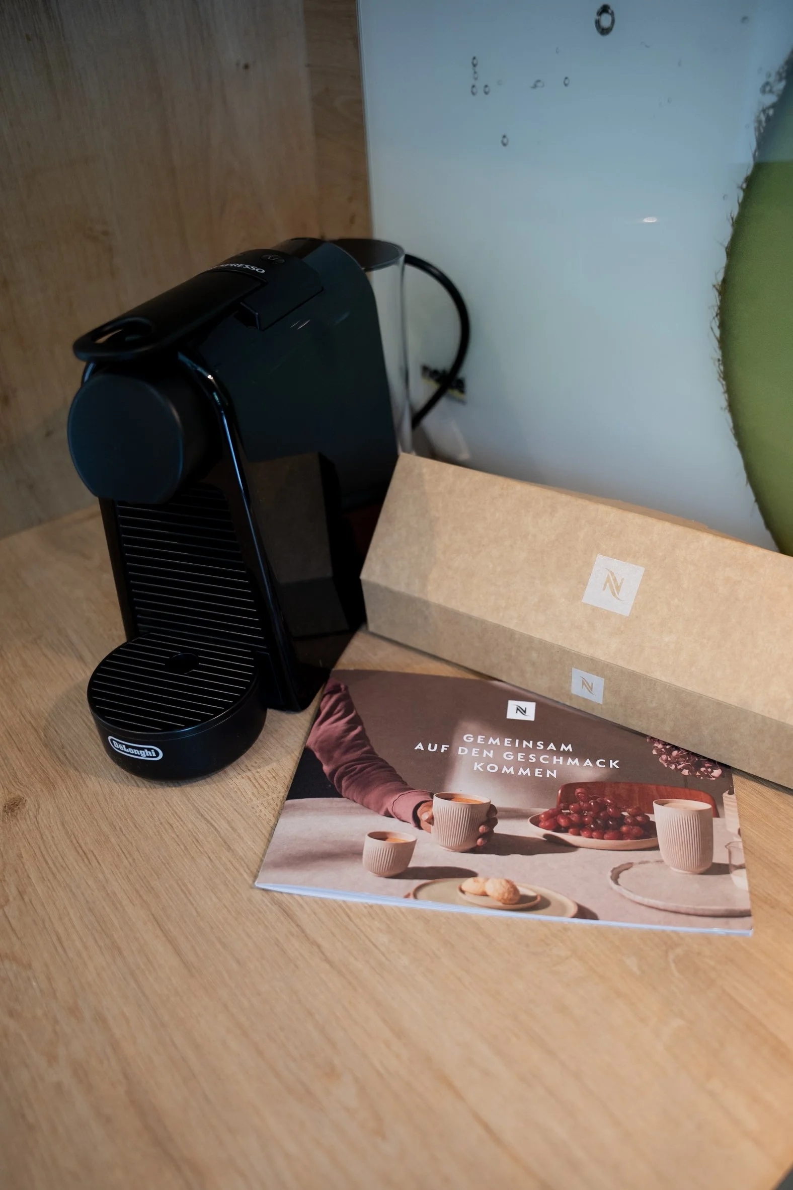 Ein Schwarz-Weiß-Kaffeemaschine auf einem Holztablett, daneben eine braune Brottasche und eine Broschüre mit der Aufschrift „Gemeinsam auf den Geschmack kommen“ und einem Bild von Tassen Kaffee.