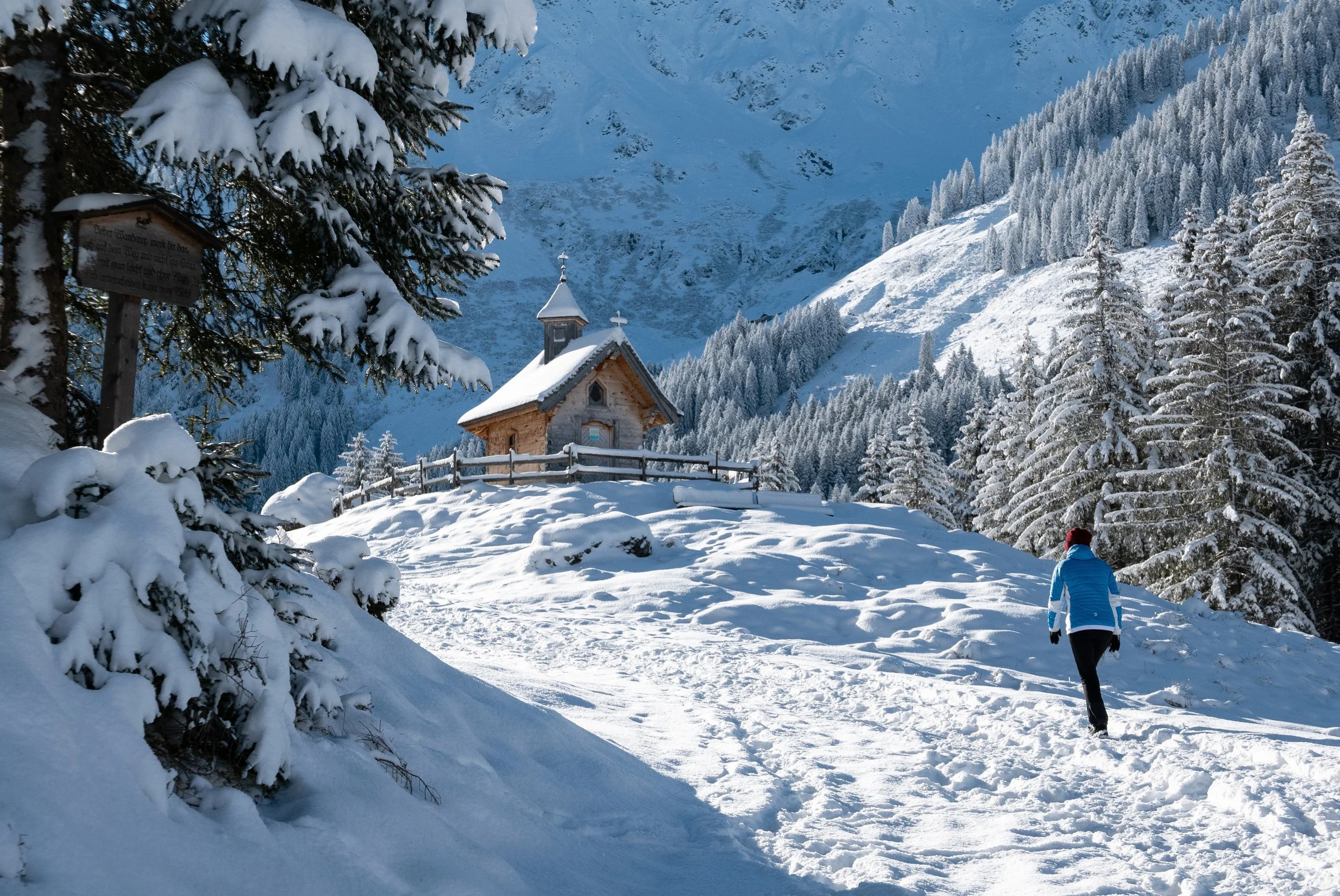 Eine verschneite Winterlandschaft mit einer kleinen Holzkapelle auf einem Hügel, umgeben von Tannenbäumen. Ein Wanderer in blauer Jacke und rotem Hut geht den verschneiten Weg entlang.