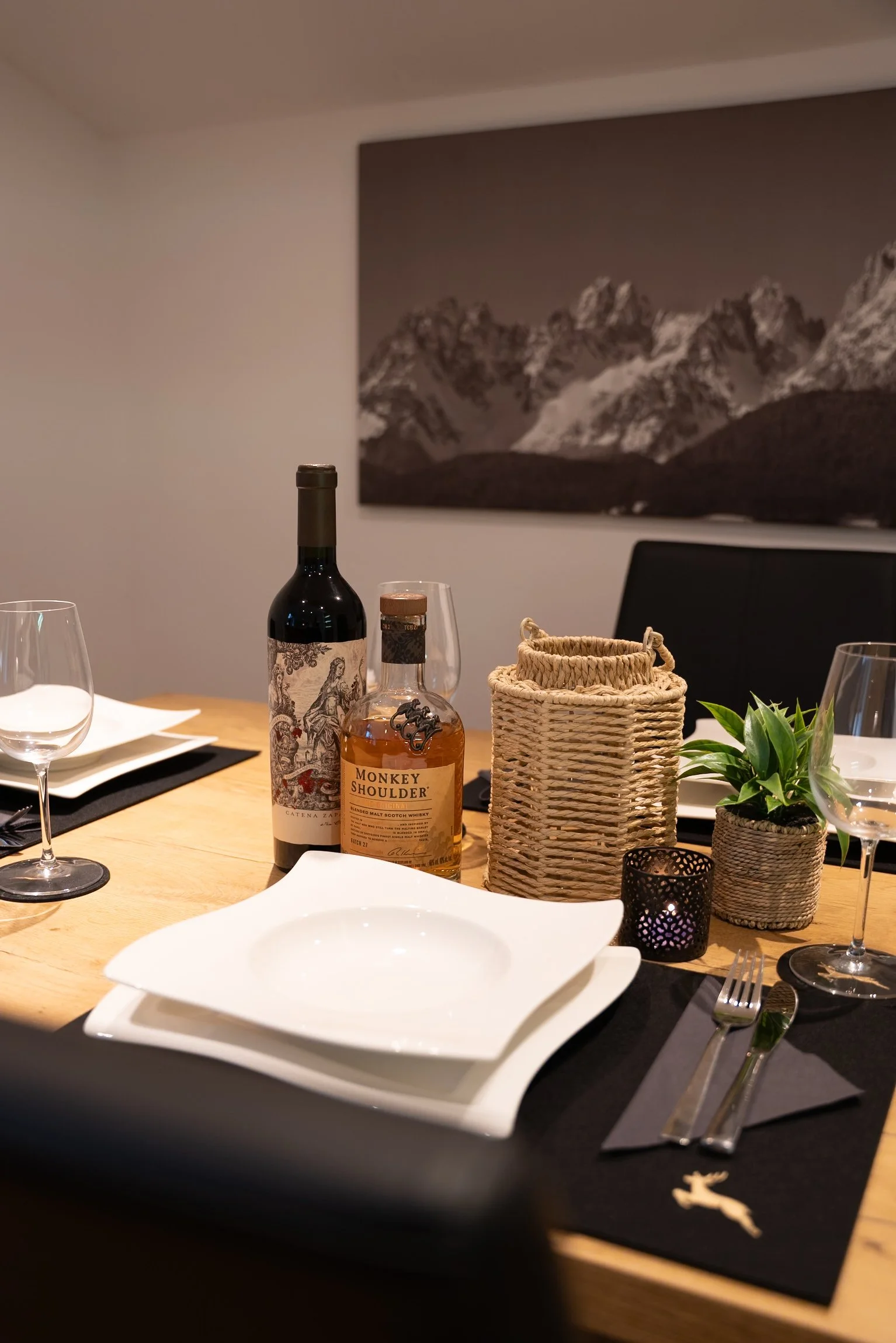 Ein gedeckter Esstisch mit weißen Tellern, Weingläsern, einer Flasche Rotwein, einem Whisky, einer kleinen Pflanze in einem Korb, einem schwarzen Teelicht und einem runden hellen Korb. Im Hintergrund ein Wandbild von Bergen.