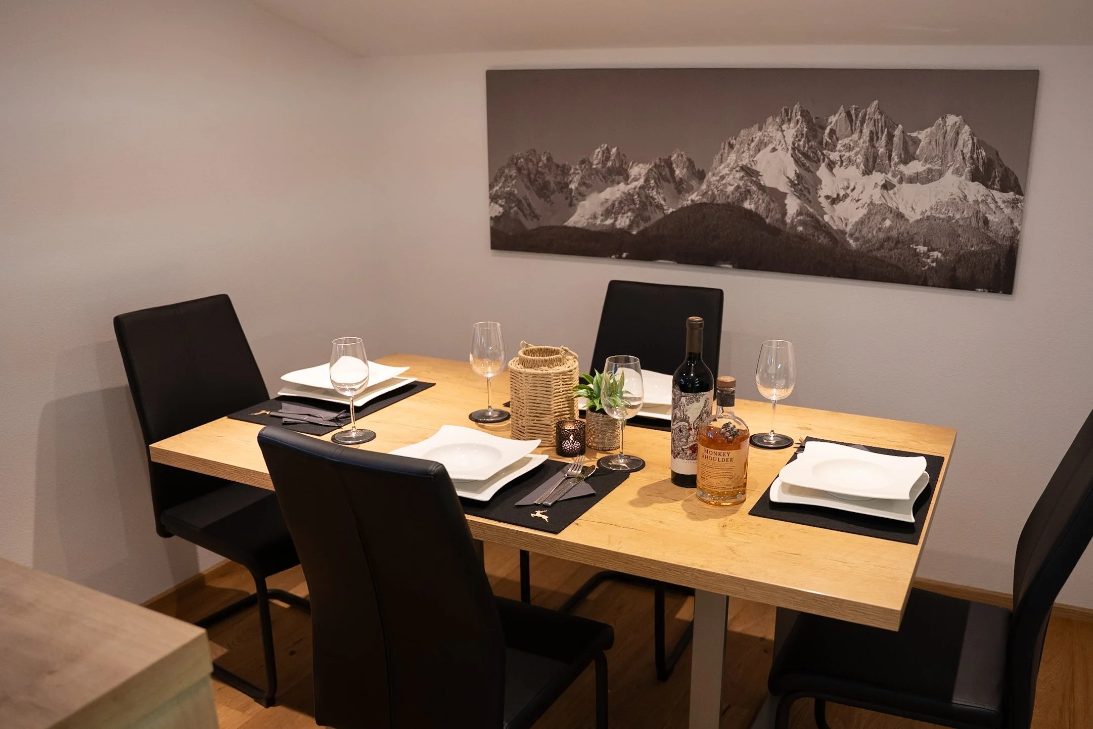 Ein gedeckter Esszimmertisch mit fünf schwarzen Stühlen, weißen Tellern, Weingläsern, Besteck, Flasche Wein, Spirituosenflasche und dekorativen Gegenständen, vor einer Wand mit Bergen im Schwarz-Weiß-Print.