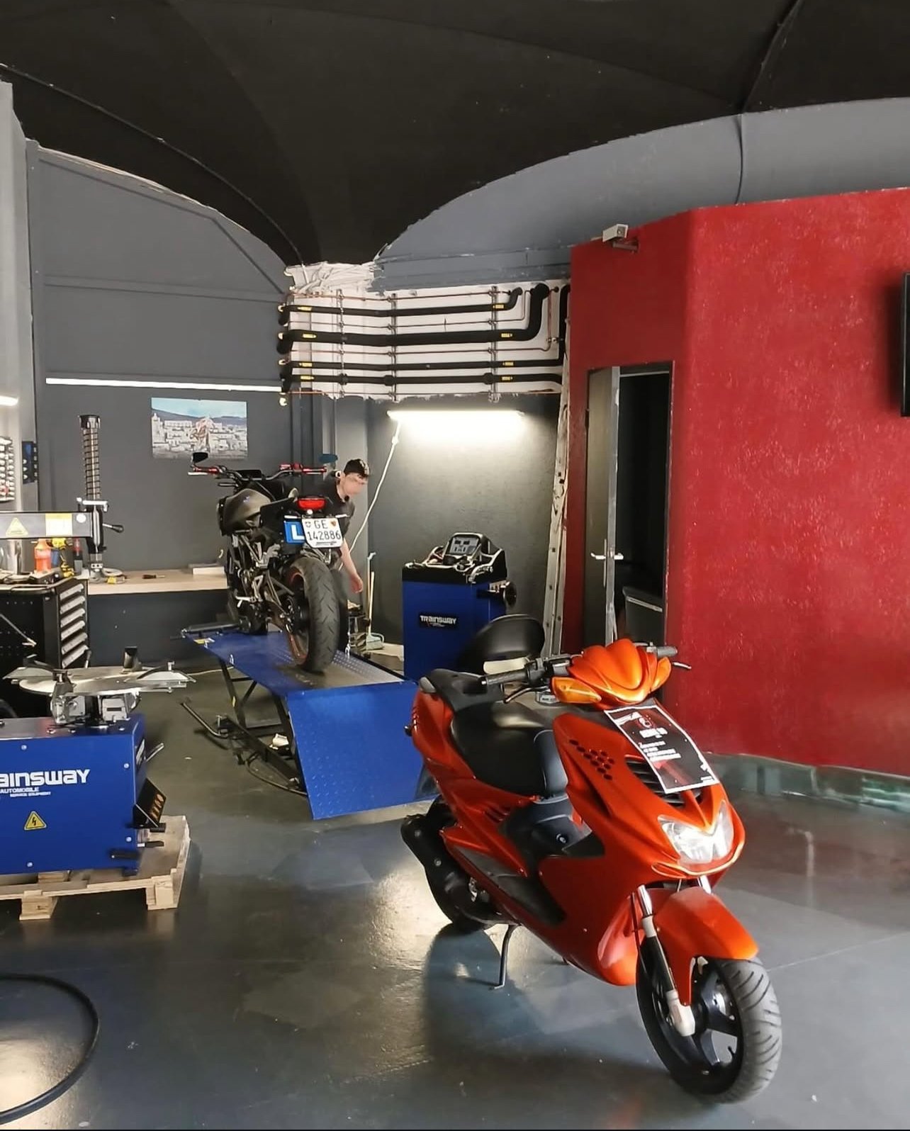 Atelier de réparation de motos avec une moto sur une plateforme de levage et un scooter rouge à l'avant.