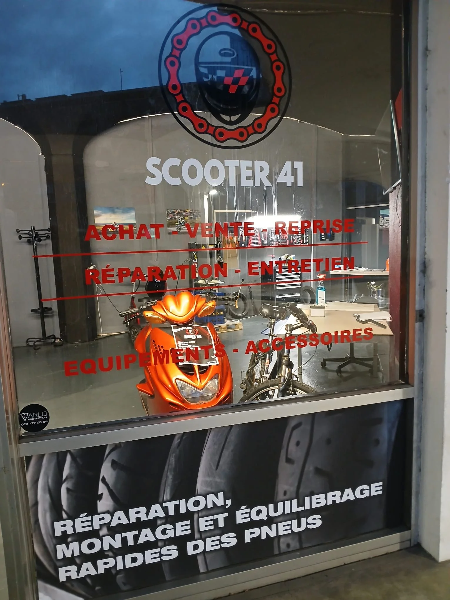 Vitrine d'un magasin de scooters avec un scooter orange à l'intérieur. Le texte sur la vitre indique "SCOOTER 41", offrant des services d'achat, vente, reprise, réparation et entretien, ainsi que des équipements et accessoires. En bas, un autre texte mentionne "Réparation, montage et équilibrage rapides des pneus".