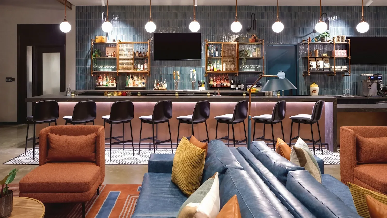 Upscale Hyatt hotel lobby bar with glowing backlit counter, black bar stools, blue tile wall, and cozy mid-century style lounge seating in orange and blue, designed for relaxed cocktails and socializing.