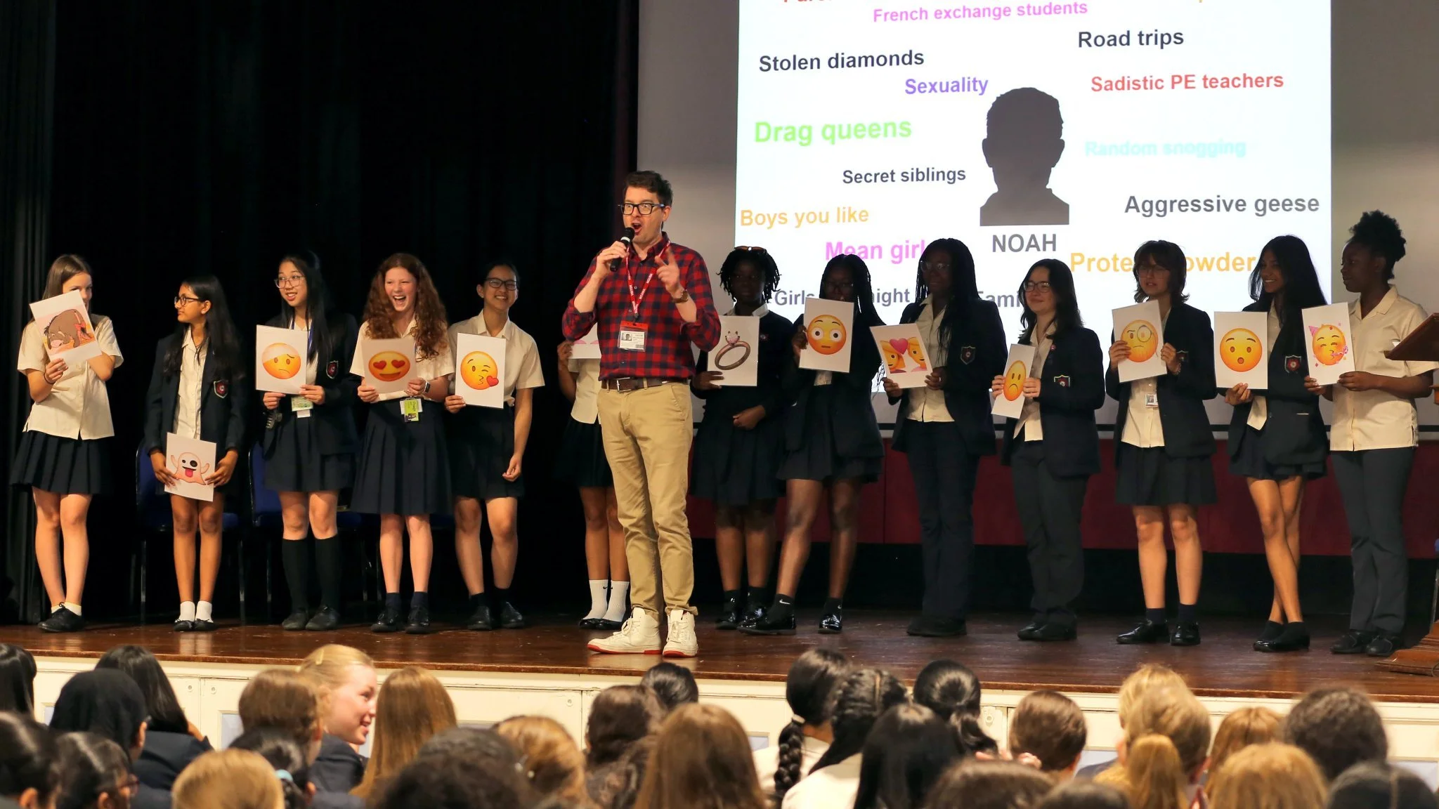 A group of students standing on stage, holding paper signs with various emoji faces. The students are wearing school uniforms and are smiling or looking at the audience. A man in a red plaid shirt and beige pants is speaking into a microphone. In the background, a presentation slide displays various words and a silhouette of a person's head.