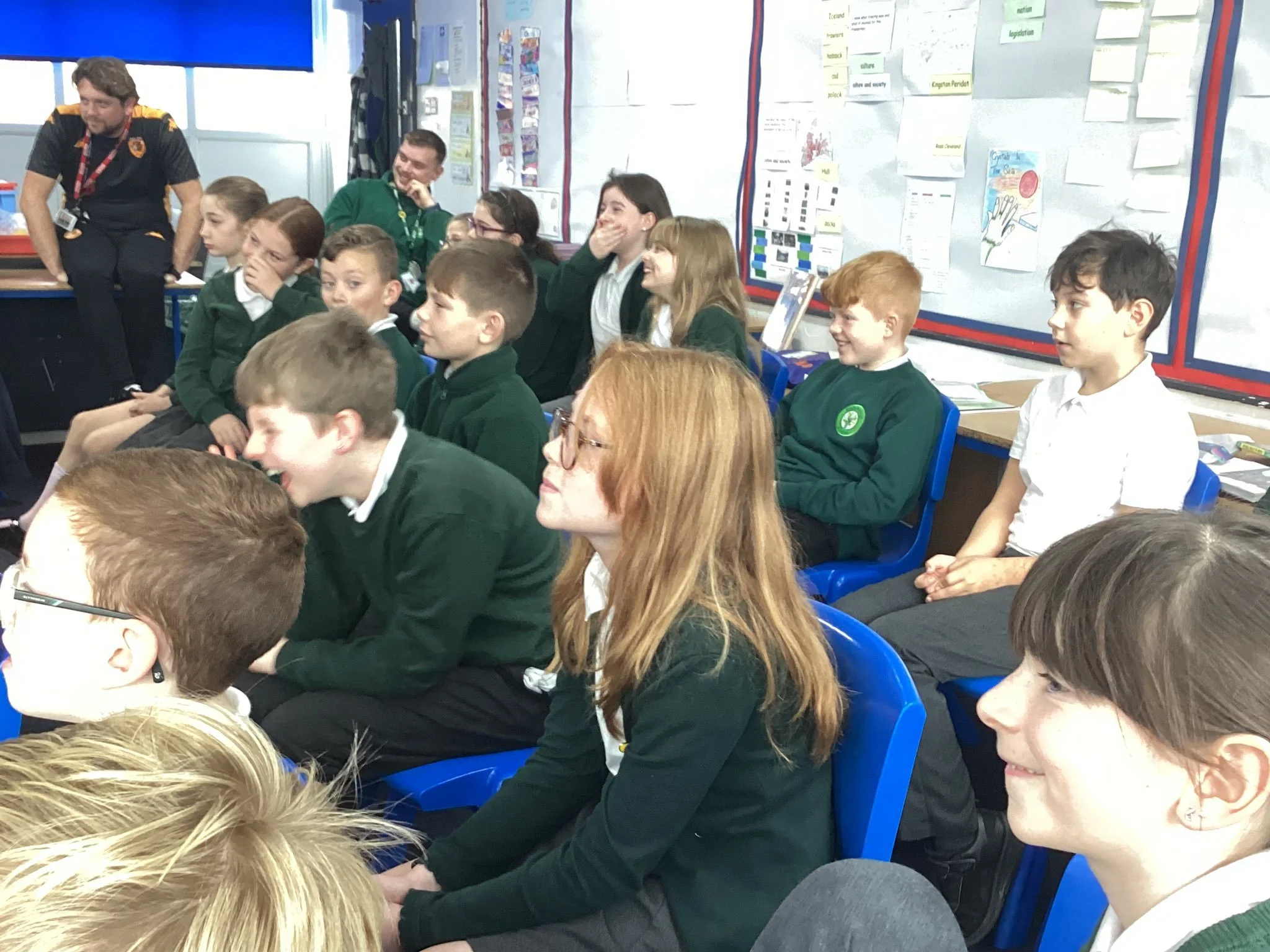 Students sitting in a classroom, listening and laughing, with colorful posters on the wall.