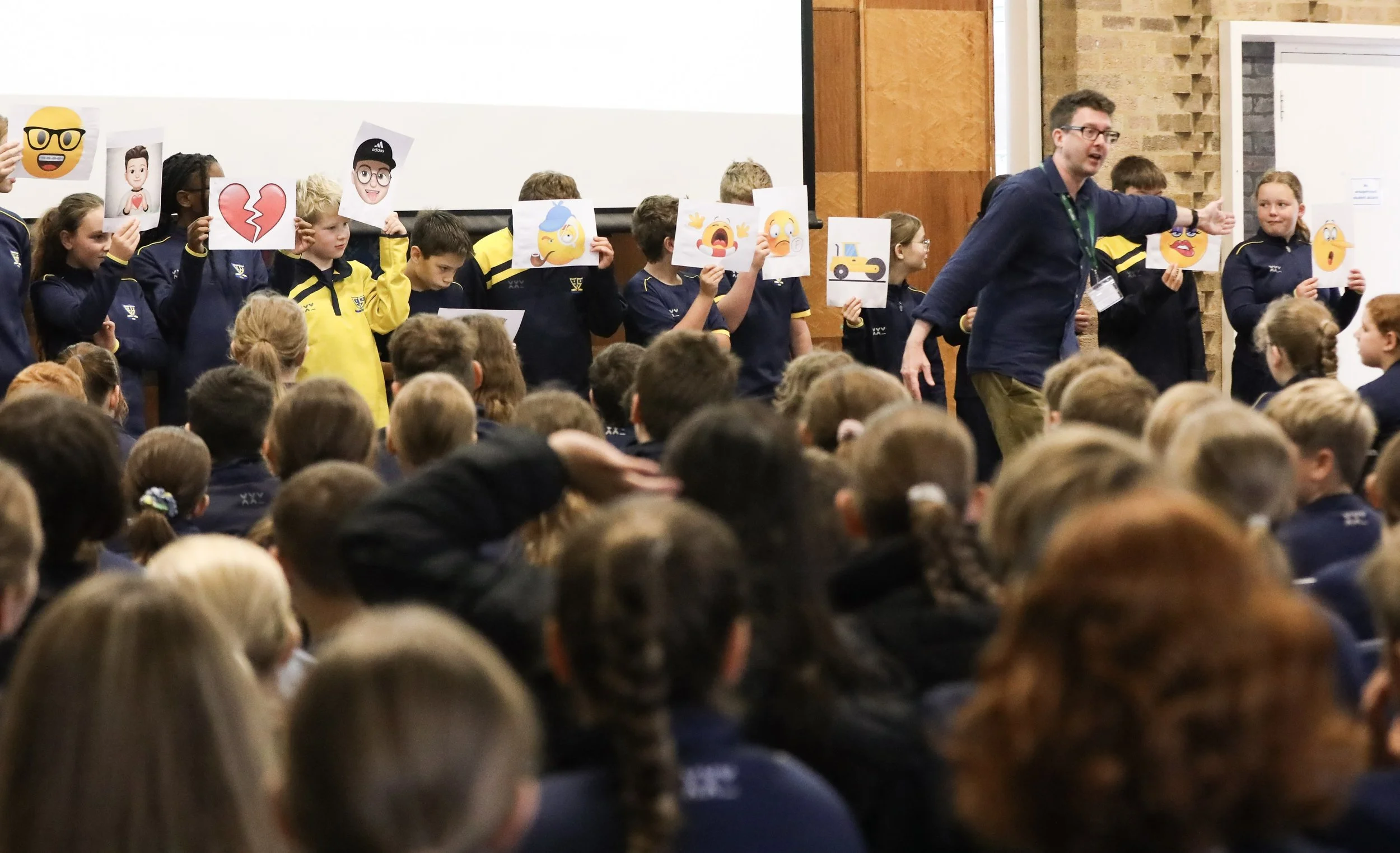 A group of children and Simon James Green are participating in an author event involving holding up large paper emojis with various expressions and objects, in front of an audience of seated children.