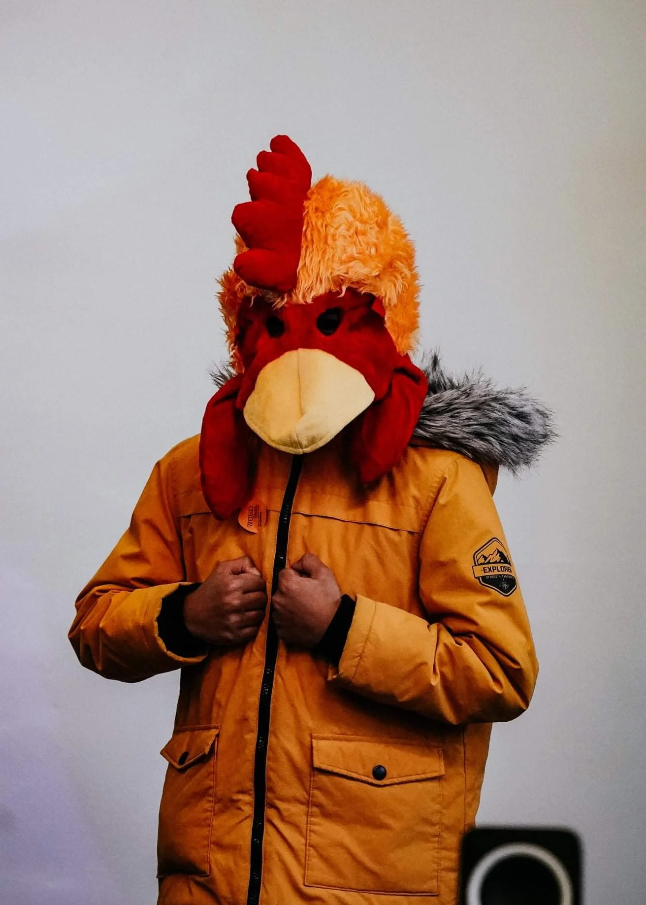 A boy wearing a yellow jacket, arms held like chicken wings, and wearing a large chicken mascot head.