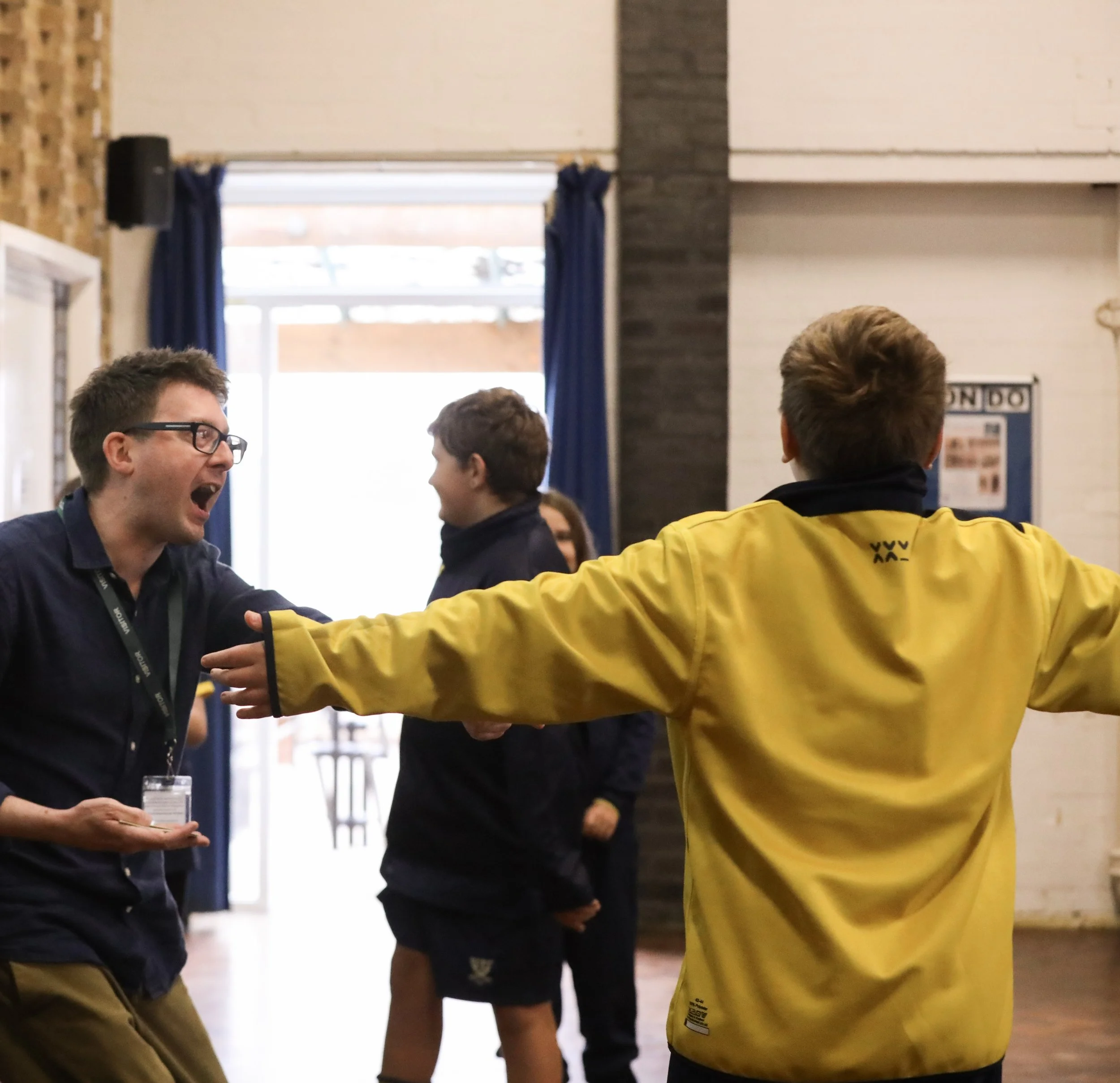 An animated scene from an author workshop, with one person wearing a yellow jacket and the other wearing a dark shirt and glasses. Other people are in the background near a door with blue curtains.