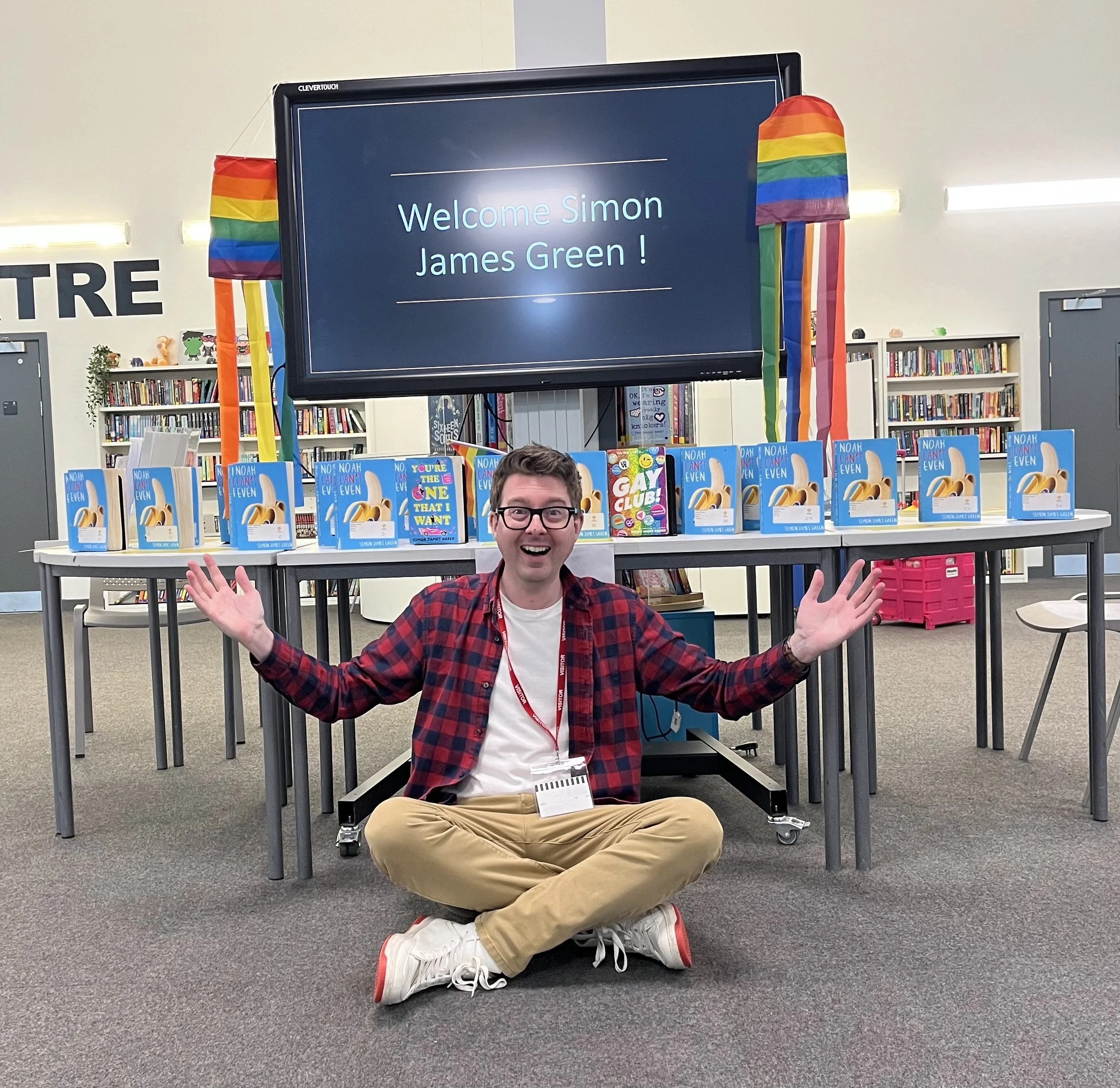 A man sitting cross-legged on the floor with arms outstretched in front of a table displaying multiple copies of a children's book titled 'Noah Can Even'. Behind him, a large screen reads 'Welcome Simon James Green!'. Rainbow-colored paper lanterns are hung on either side of the screen, and the setting appears to be a library.