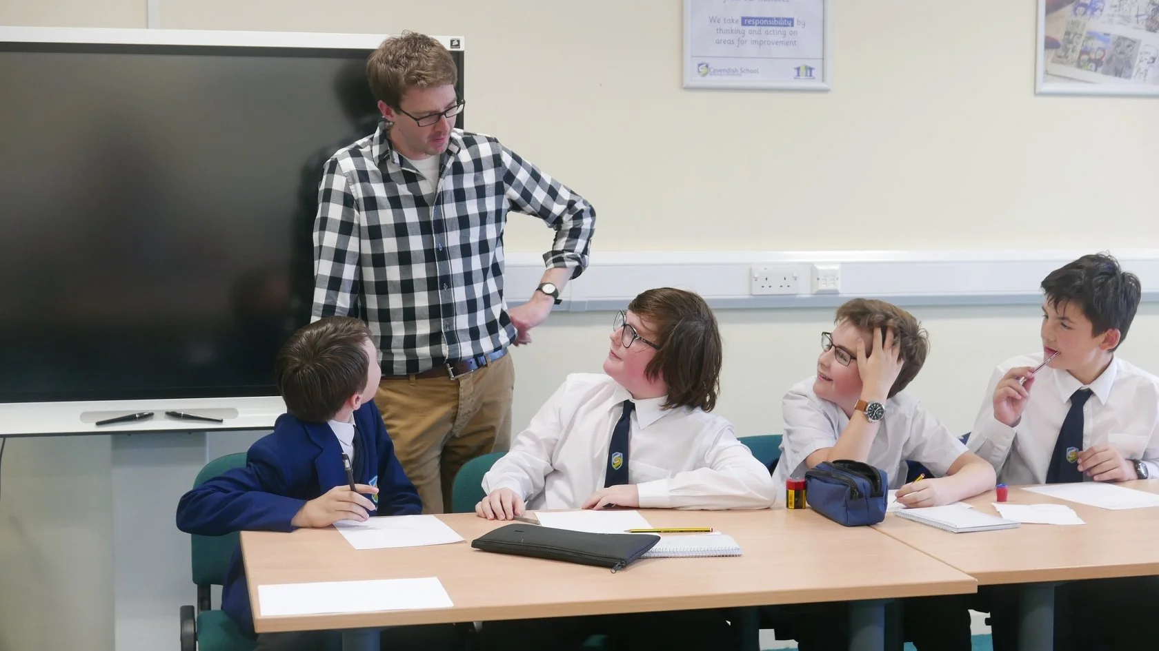 Simon standing and talking to four students seated at a classroom table. The students are wearing school uniforms and are engaged in conversation with the Simon.