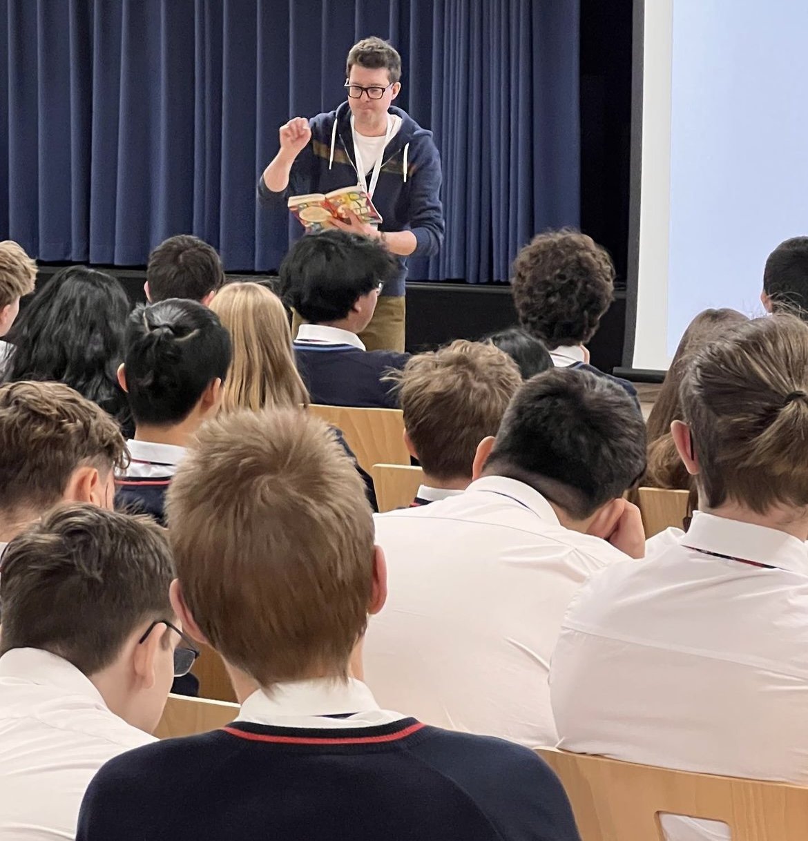 Simon James Green reading from one of his books to a seated audience of students in a school auditorium with a blue curtain in the background.