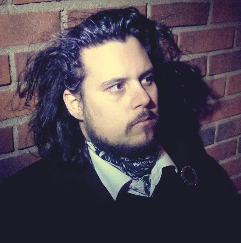 Man med långt, mörkt hår och skägg, klädd i svart och vitt, står mot en röd tegelvägg.