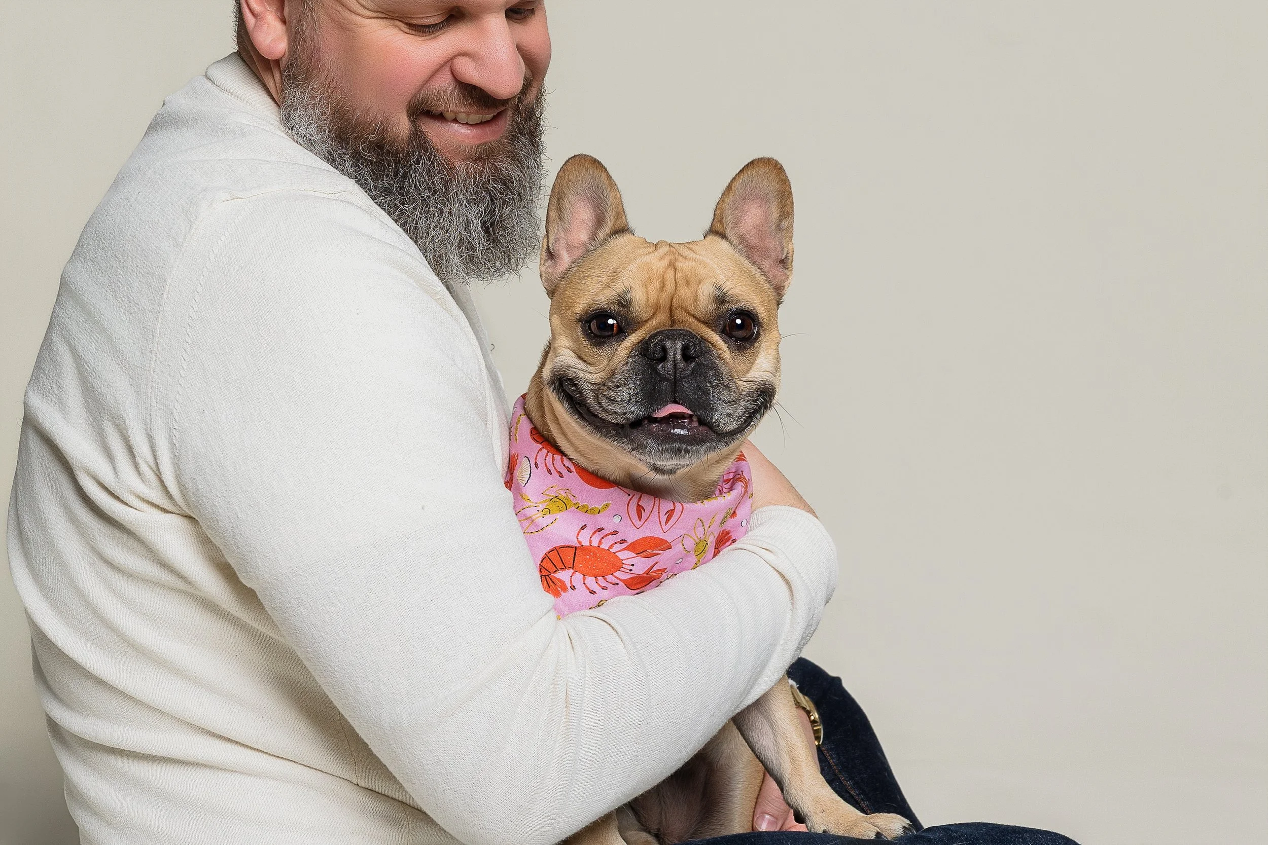A man with a gray beard smiling while holding a smiling French Bulldog wearing a pink bandana with orange designs against a plain background.