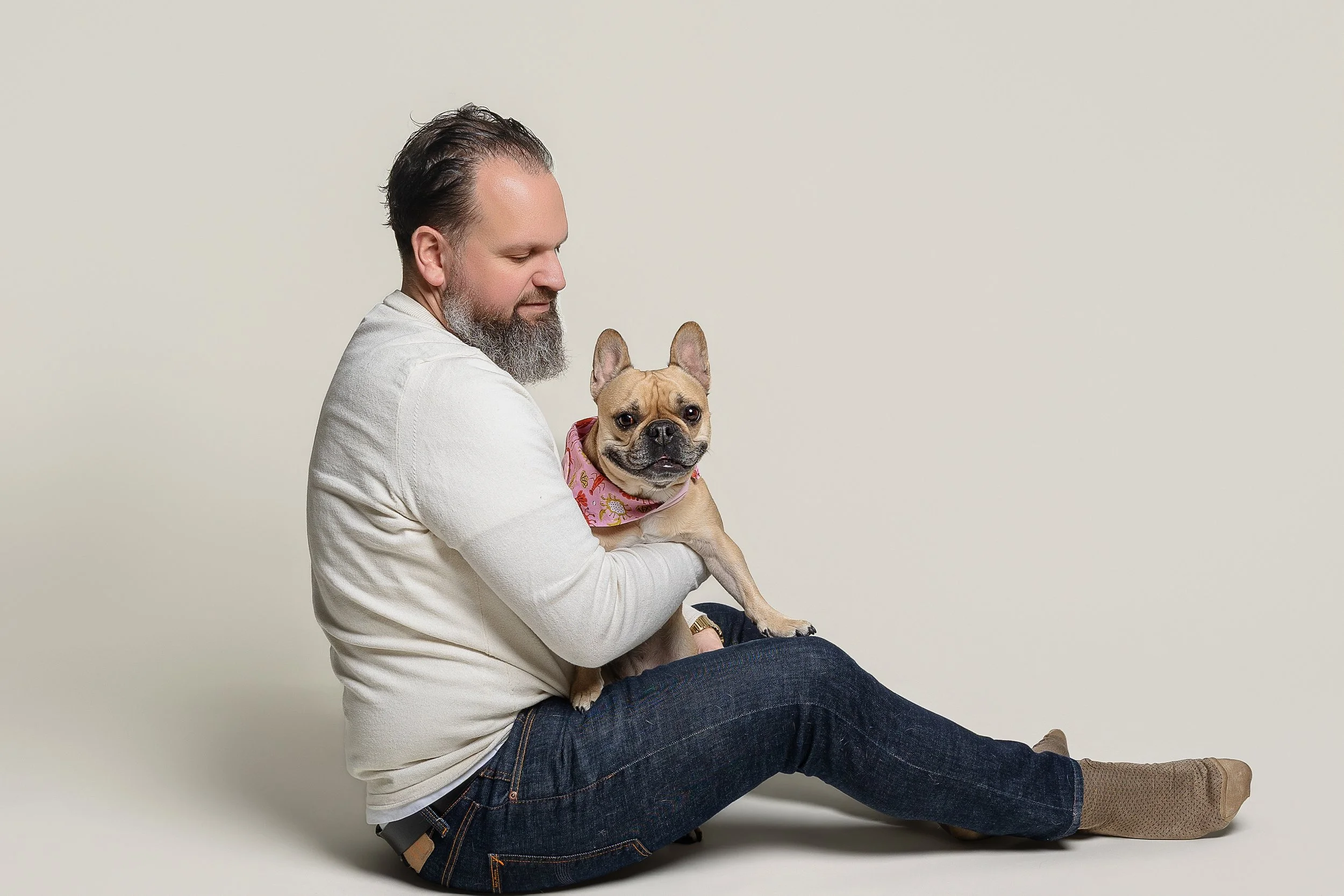 A man with a beard and wearing a white sweater and jeans sitting on the floor holding a French Bulldog dog with a pink bandana against a plain beige background.