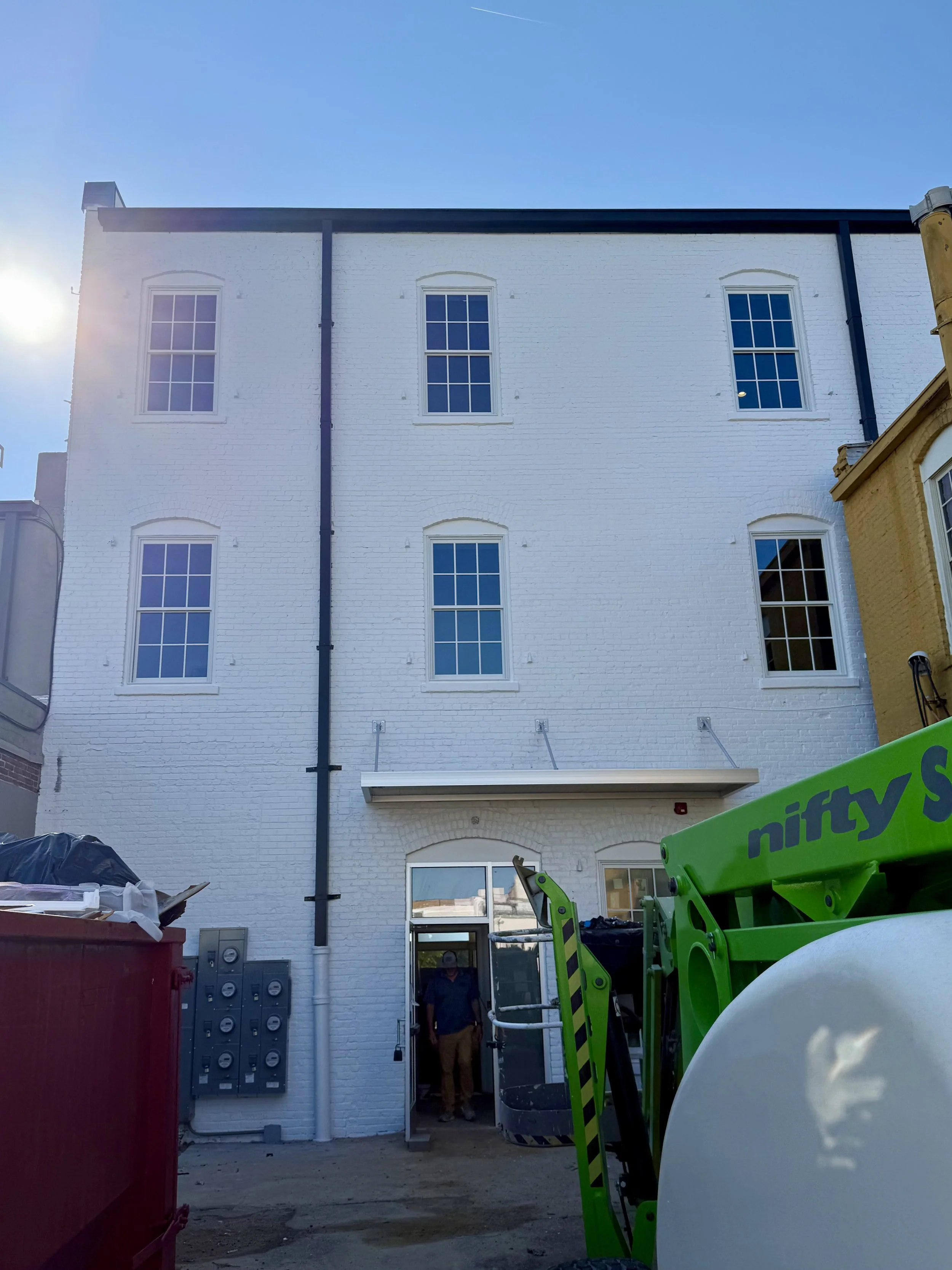 The back of a white brick building with four windows, a door, and a construction worker standing in the doorway. Construction equipment is visible in the foreground.