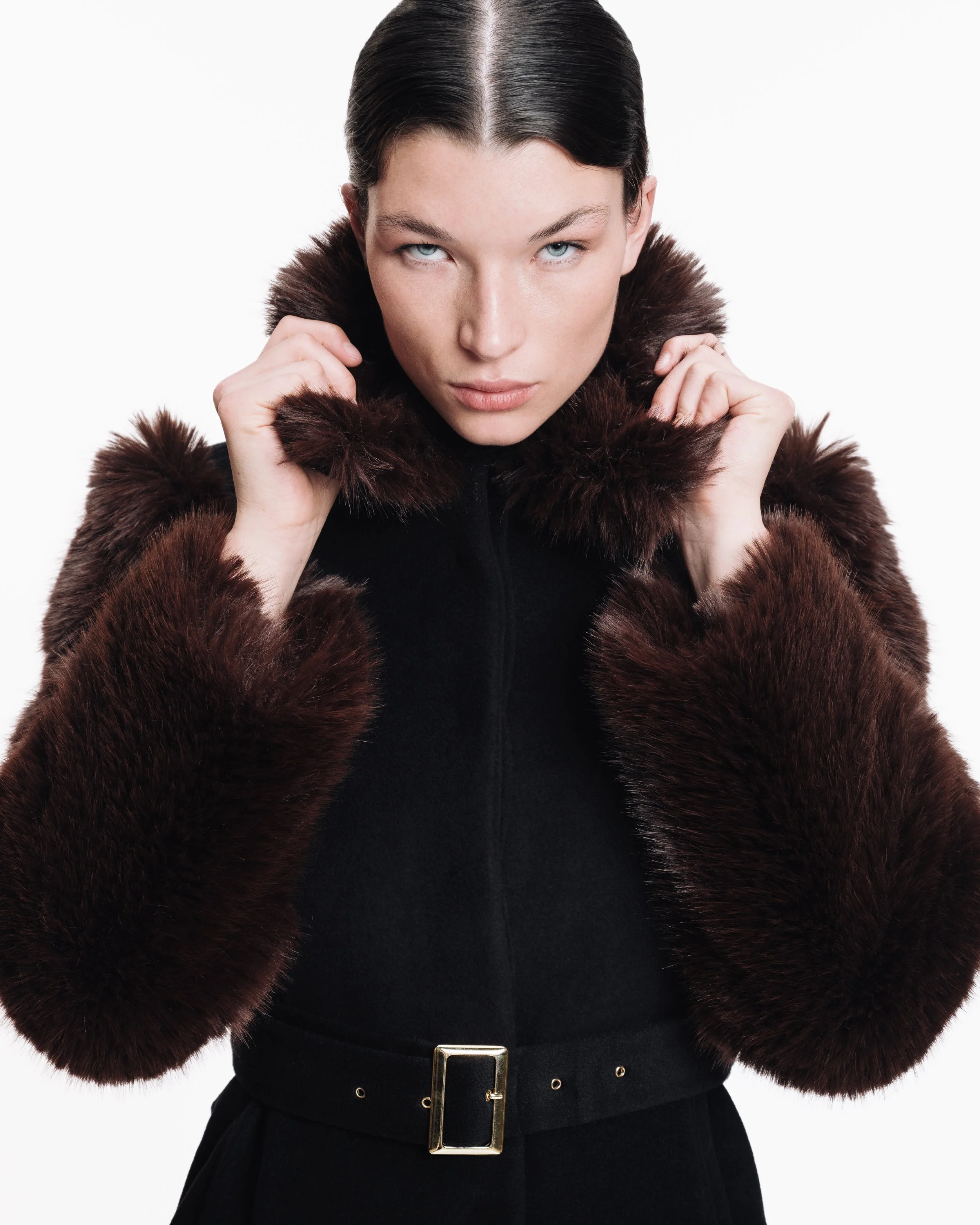 Woman wearing a black coat with dark brown fur collar and cuffs, posing with hands near her face against a plain white background.