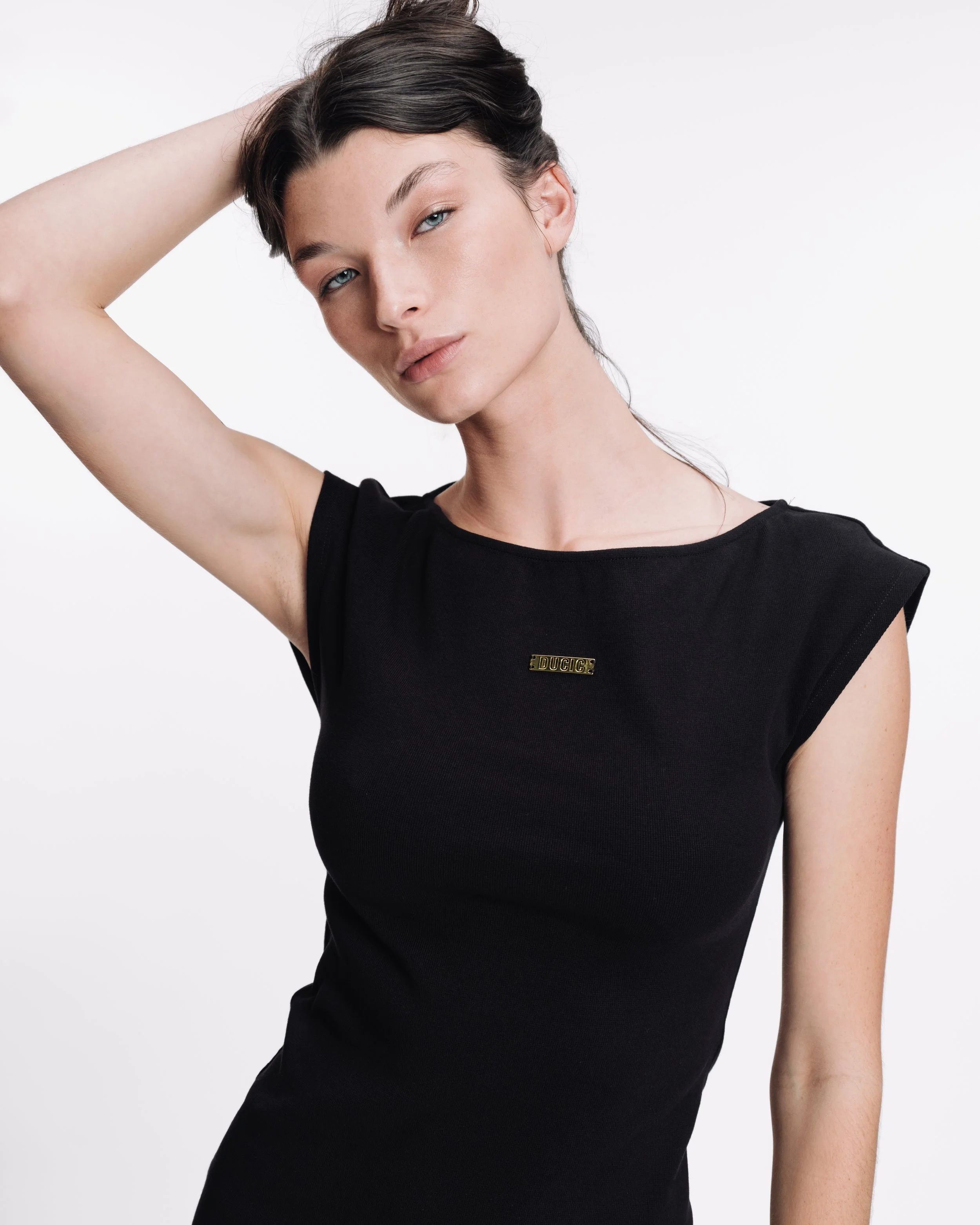 A young woman with dark hair and fair skin, wearing a black sleeveless top with a small gold pin that says 'DUCIC' on the chest, touching her hair with her left hand against a plain white background.