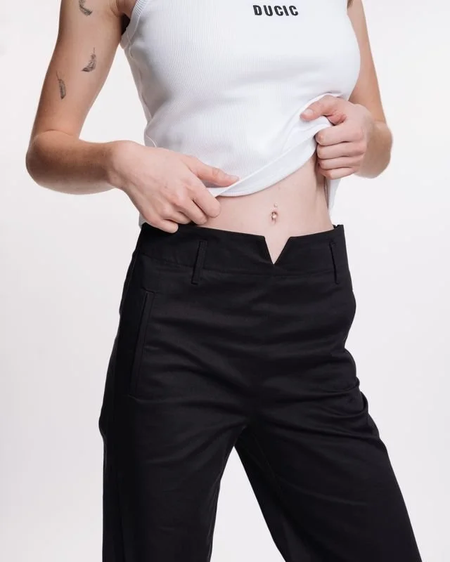 Person in a white tank top and black pants lifting their shirt to reveal their abdomen with a belly button piercing.