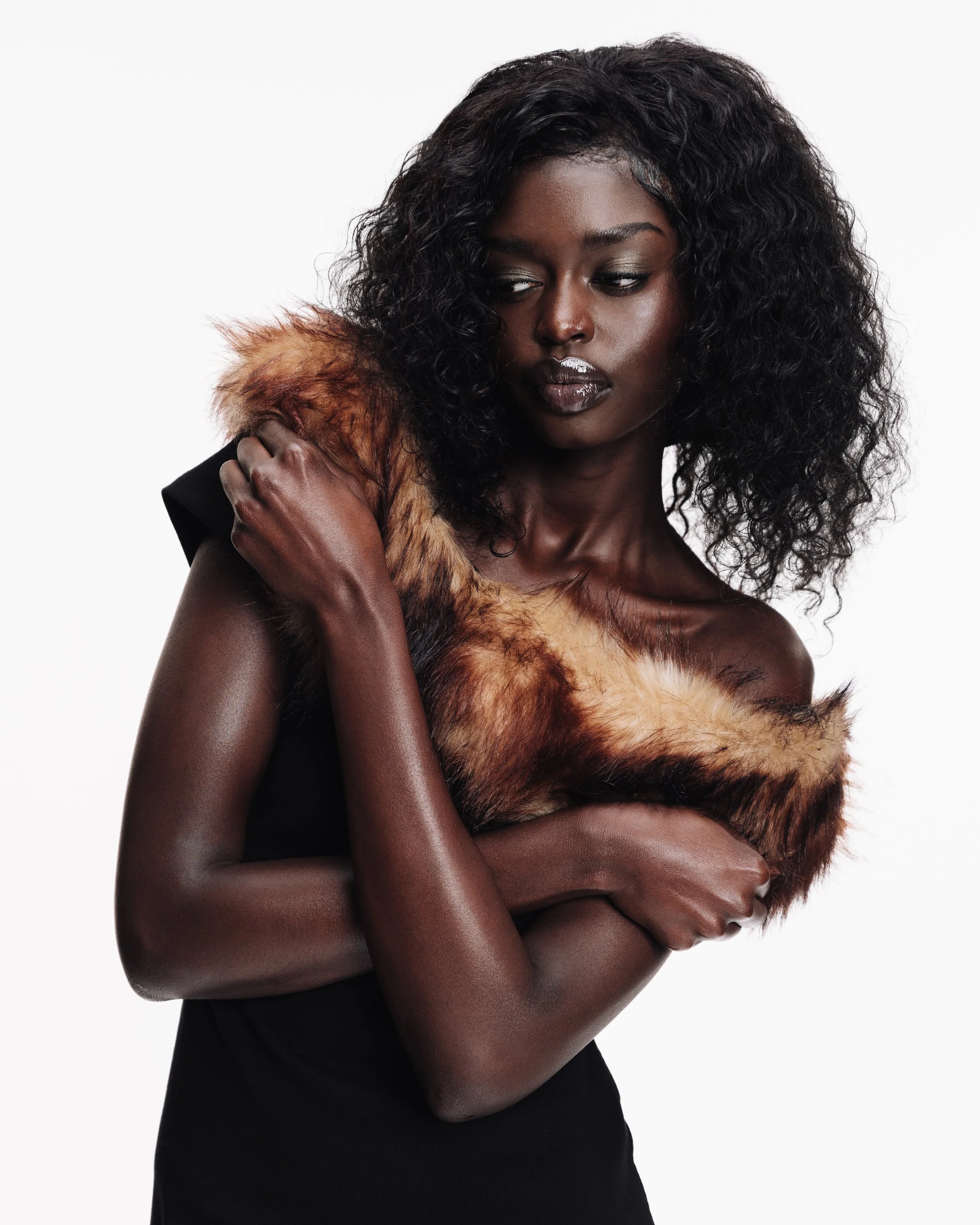 A woman with dark skin and curly black hair holding a fur stole over her shoulder, wearing a sleeveless black dress, looking downward against a plain white background.