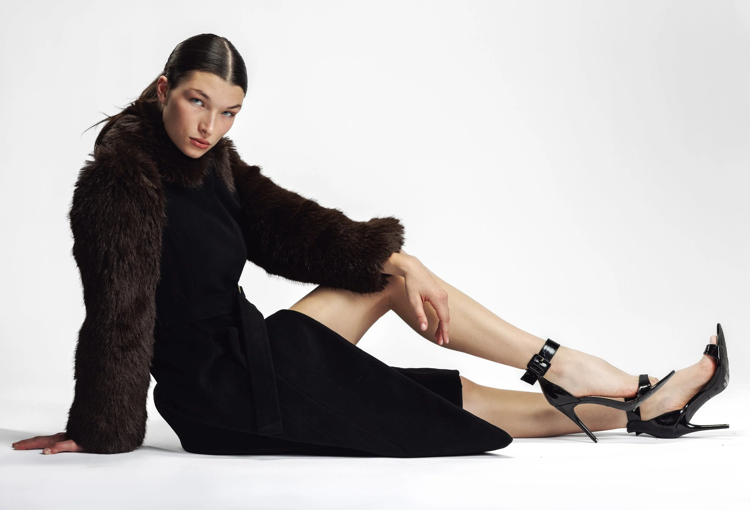 A woman with dark hair in a sleek style, wearing a black coat with fur sleeves, black high heels with ankle straps, sitting on the floor against a plain white background.