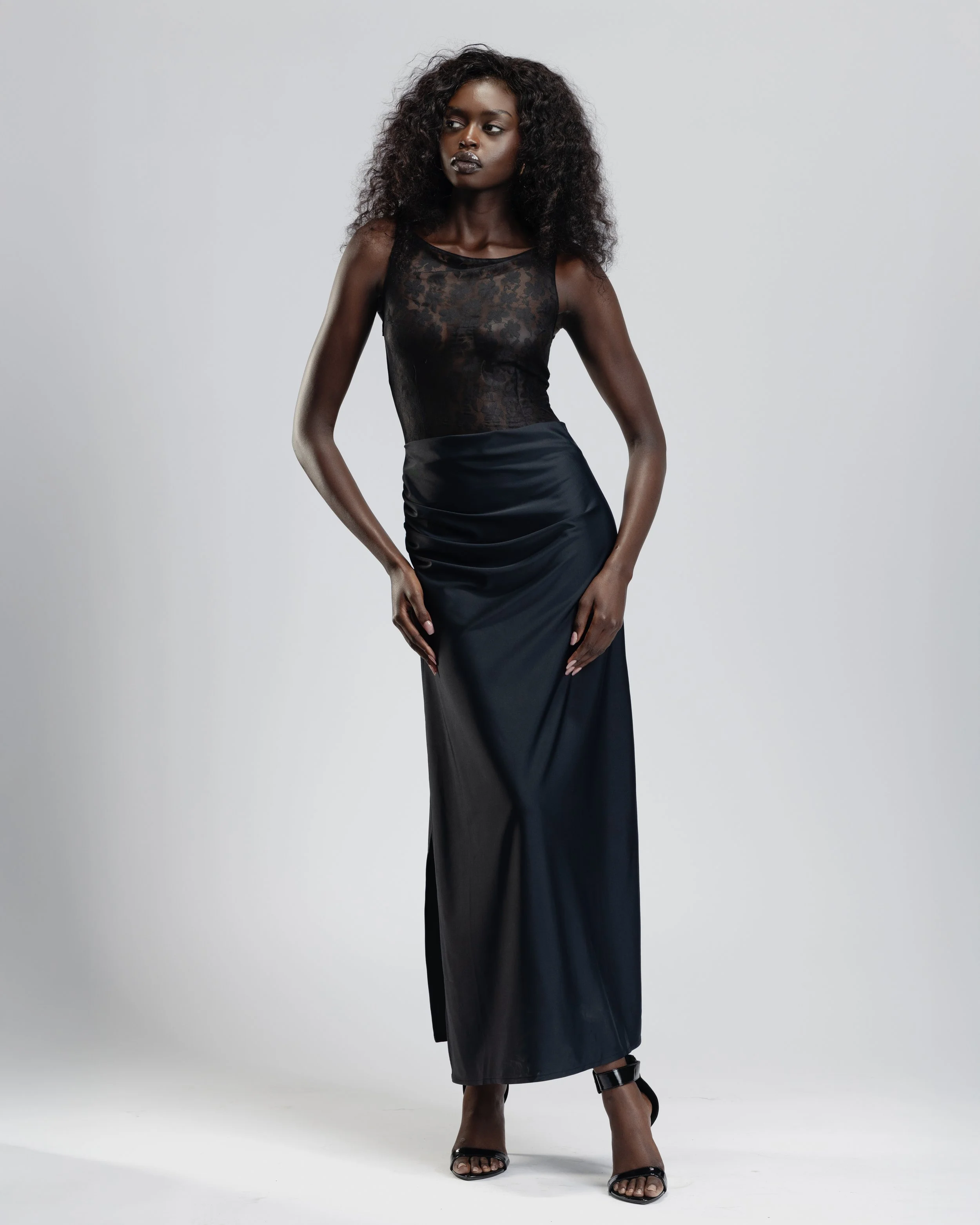 A woman with dark skin tone and curly hair wearing a black sleeveless dress with a sheer lace top and a long satin skirt, standing against a plain light gray background.
