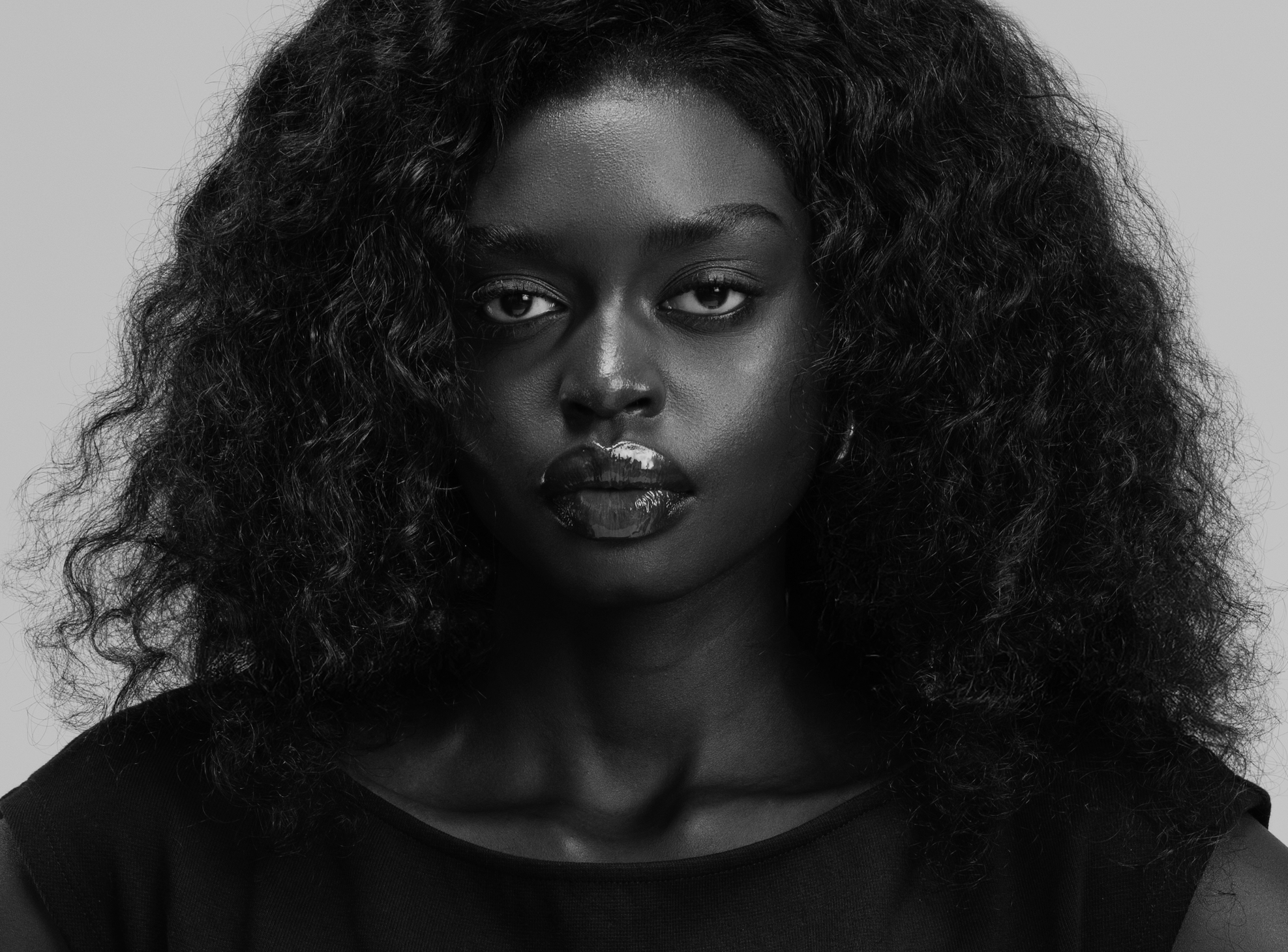 A black-and-white portrait of a woman with voluminous, curly hair and dark lipstick, looking directly at the camera.