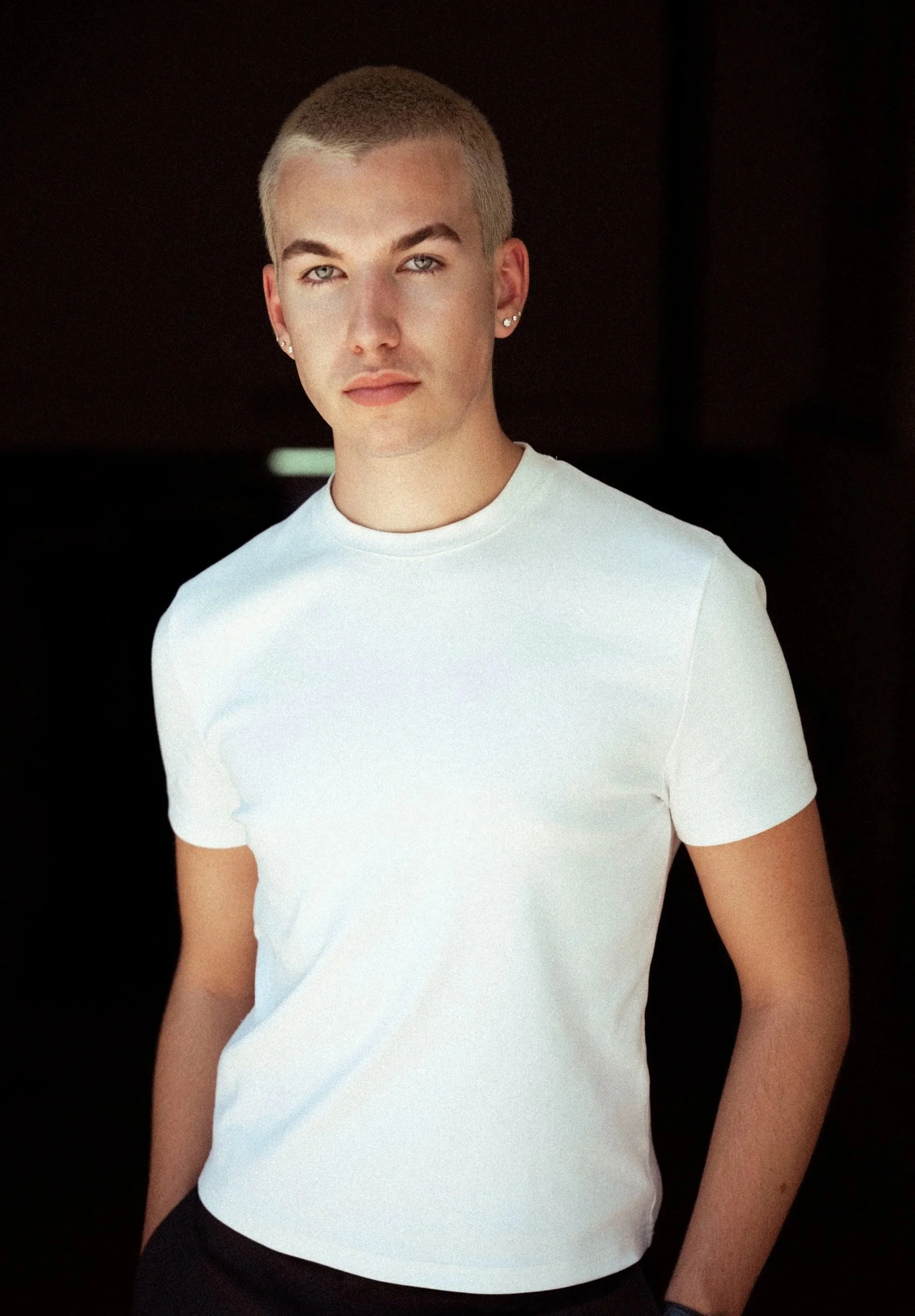 A young man with short blond hair, wearing earrings, a white t-shirt, and black pants, posing against a dark background.