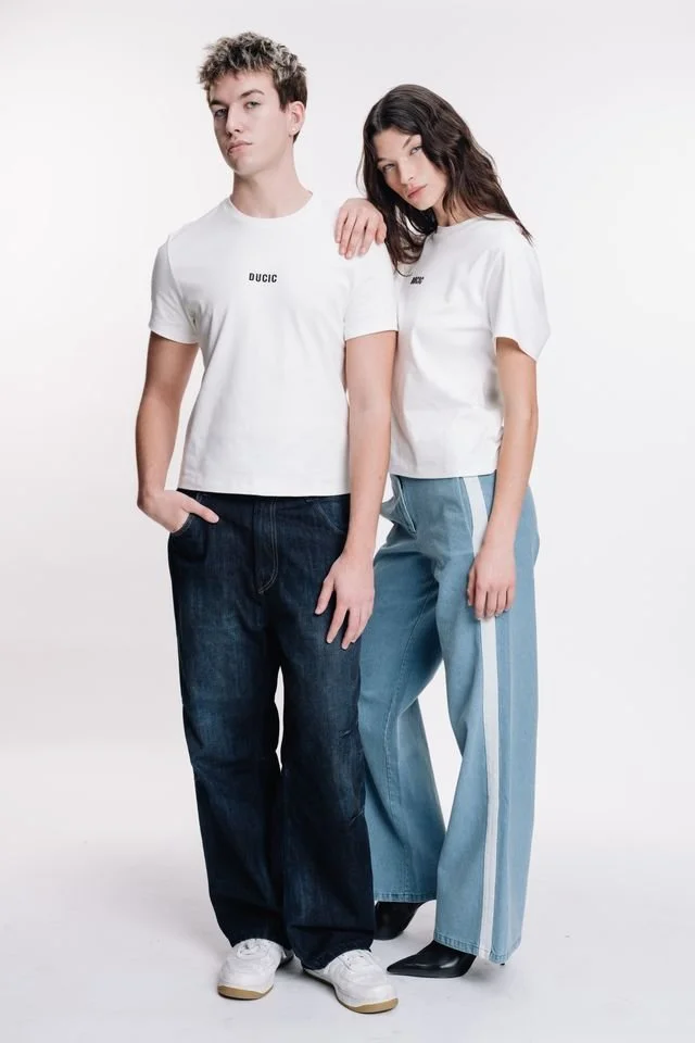 A young man and woman standing together against a white background. The man is wearing a white T-shirt and dark jeans, while the woman is wearing a white T-shirt and light blue wide-leg jeans with white stripes. They are both posing casually.