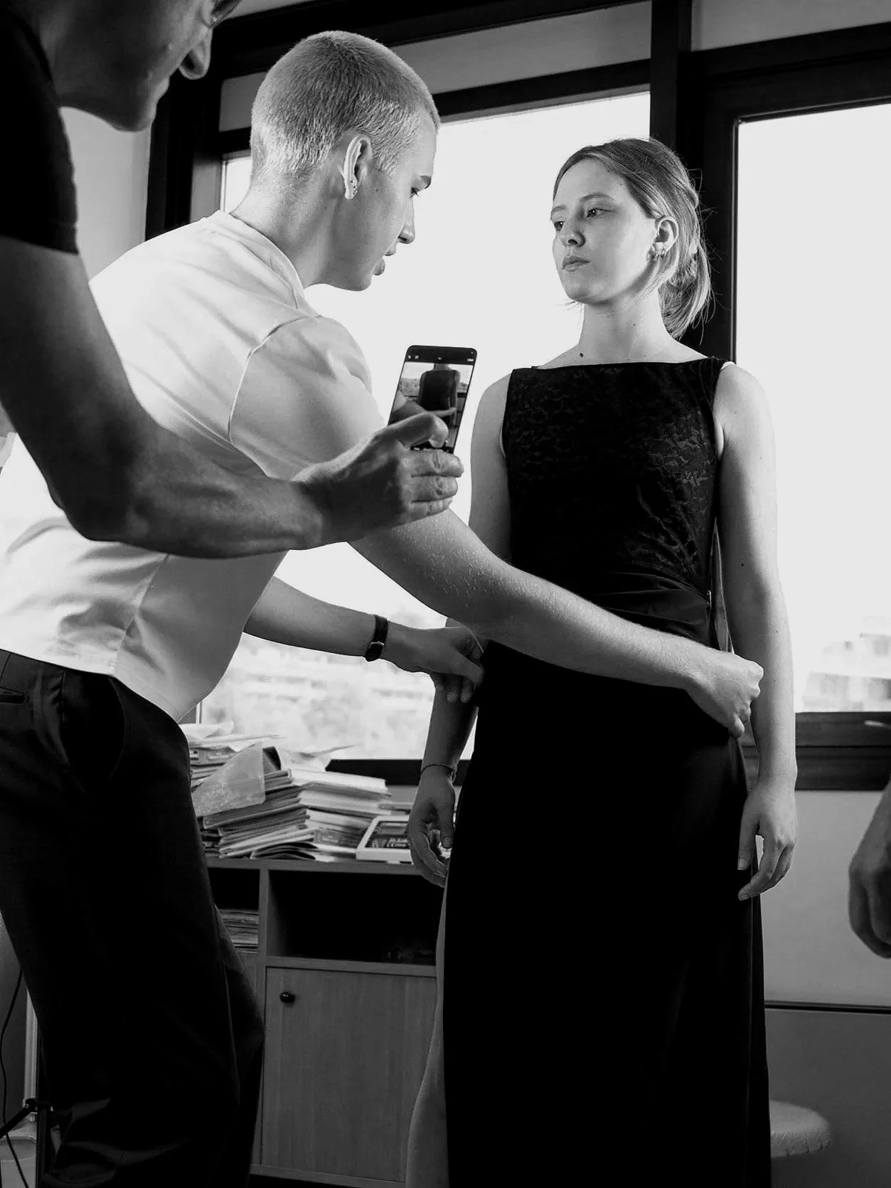 Black and white photo of a woman in a black dress with a tattoo on her arm, being photographed by a man using a smartphone, with a third person partially visible on the left side, inside an office with a large window and a desk with papers.