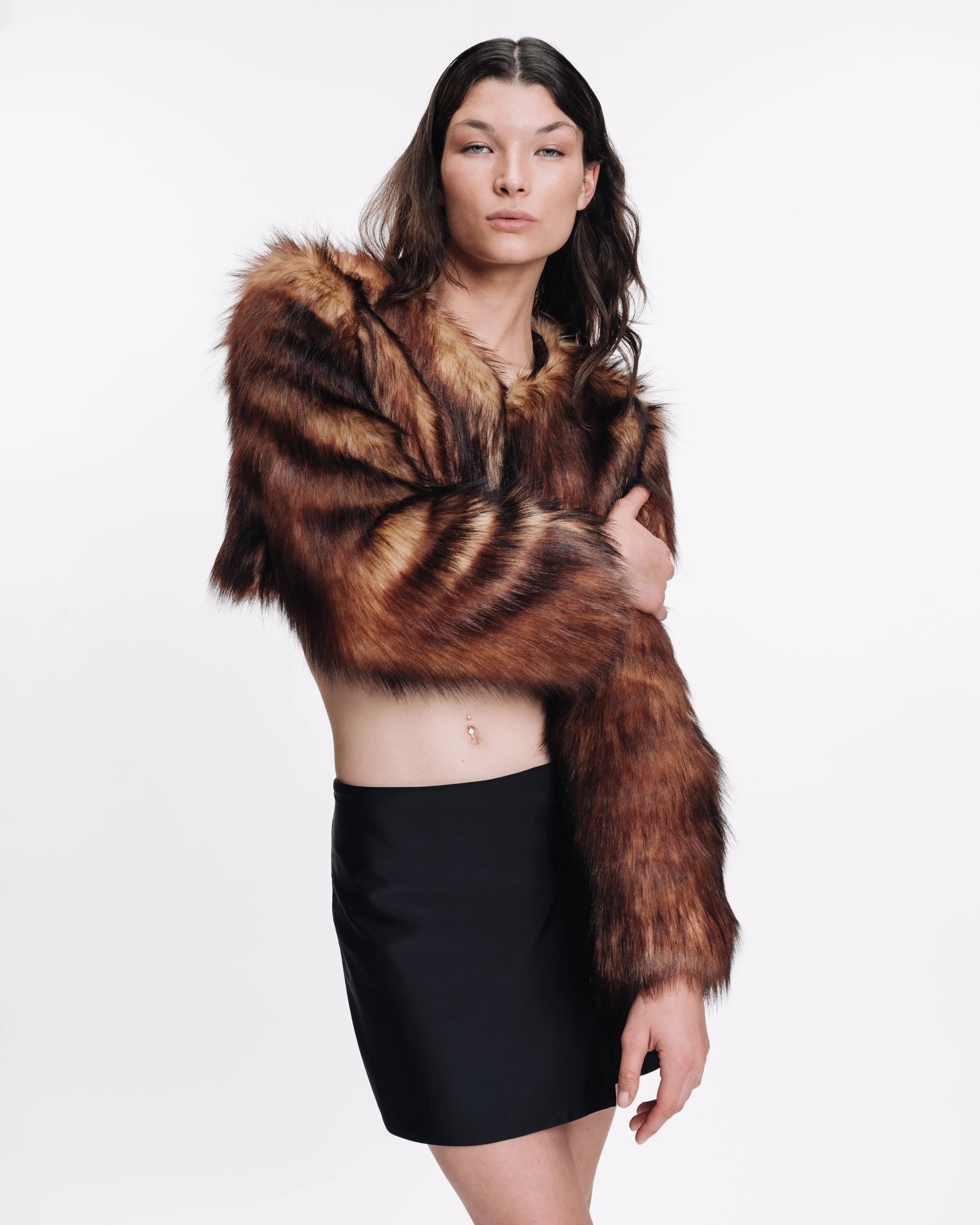 A woman with long dark hair wearing a brown fur coat and a black skirt, with a belly button piercing, stands against a plain white background.