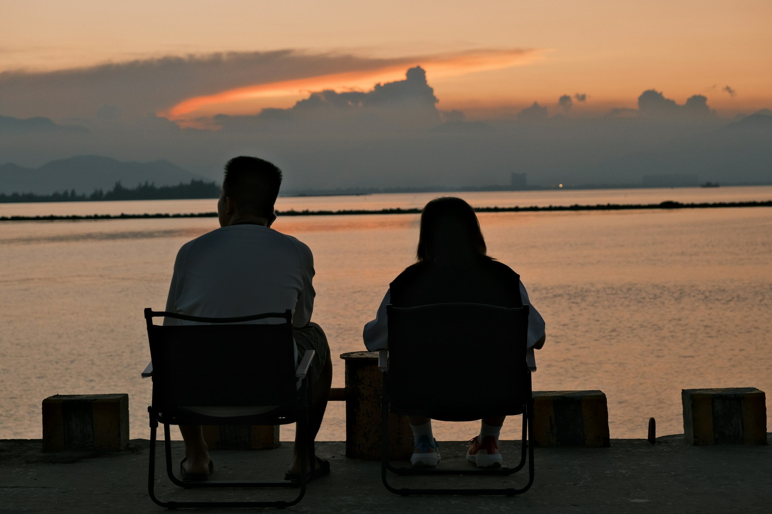 Two people are sitting on chairs by the water at sunset, facing away from the camera, with mountains in the distance and colorful sky.