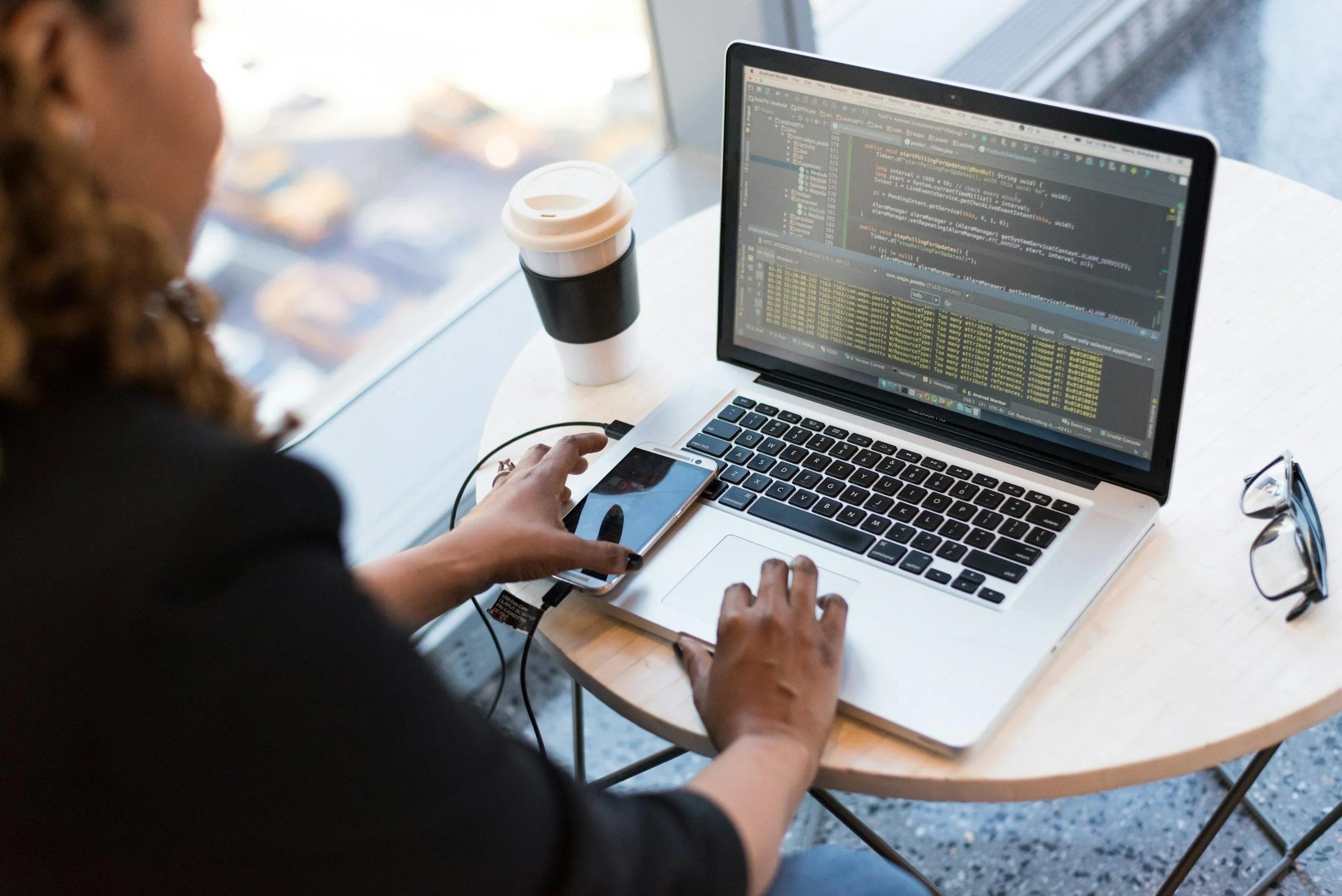 Person working on laptop with code on screen, sitting at round table near window, coffee to-go cup, glasses, and smartphone nearby.