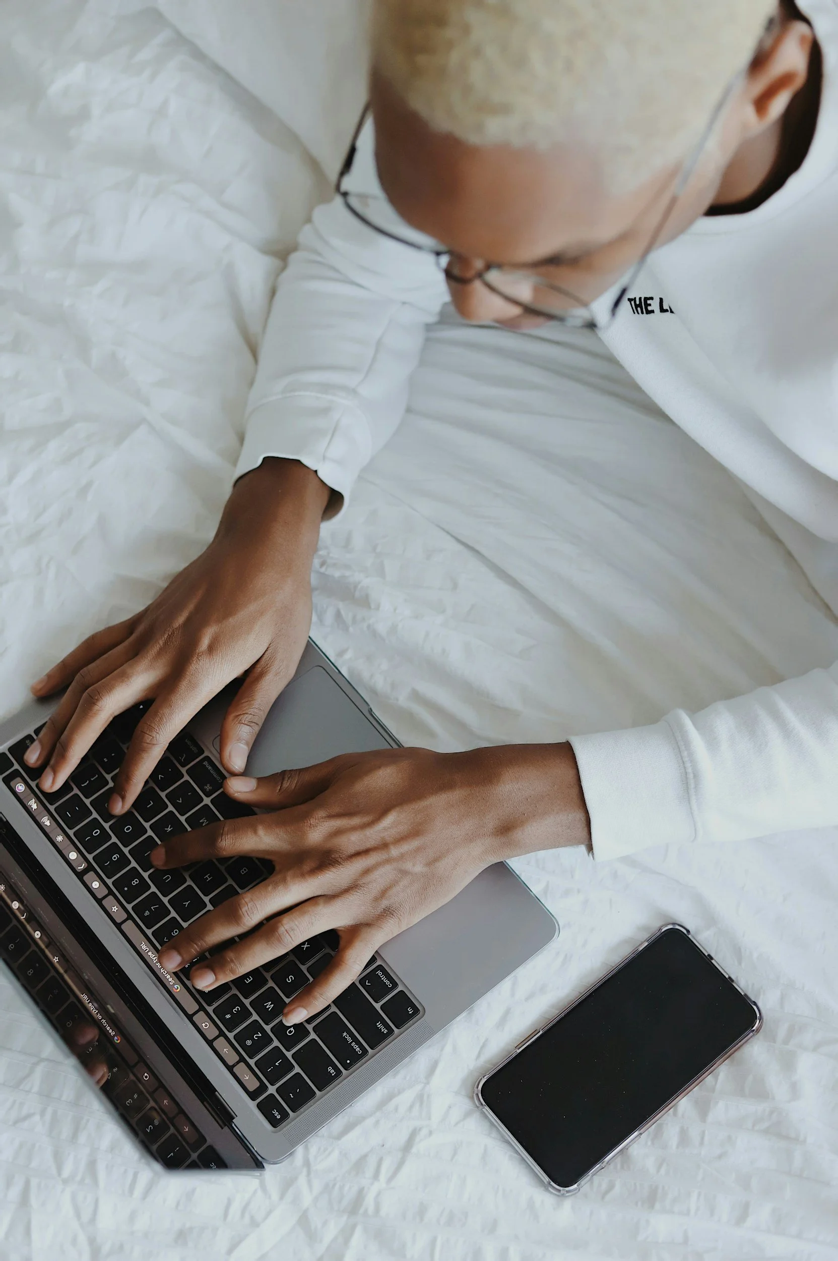 Person wearing glasses and a white shirt typing on a laptop beside a smartphone on a bed