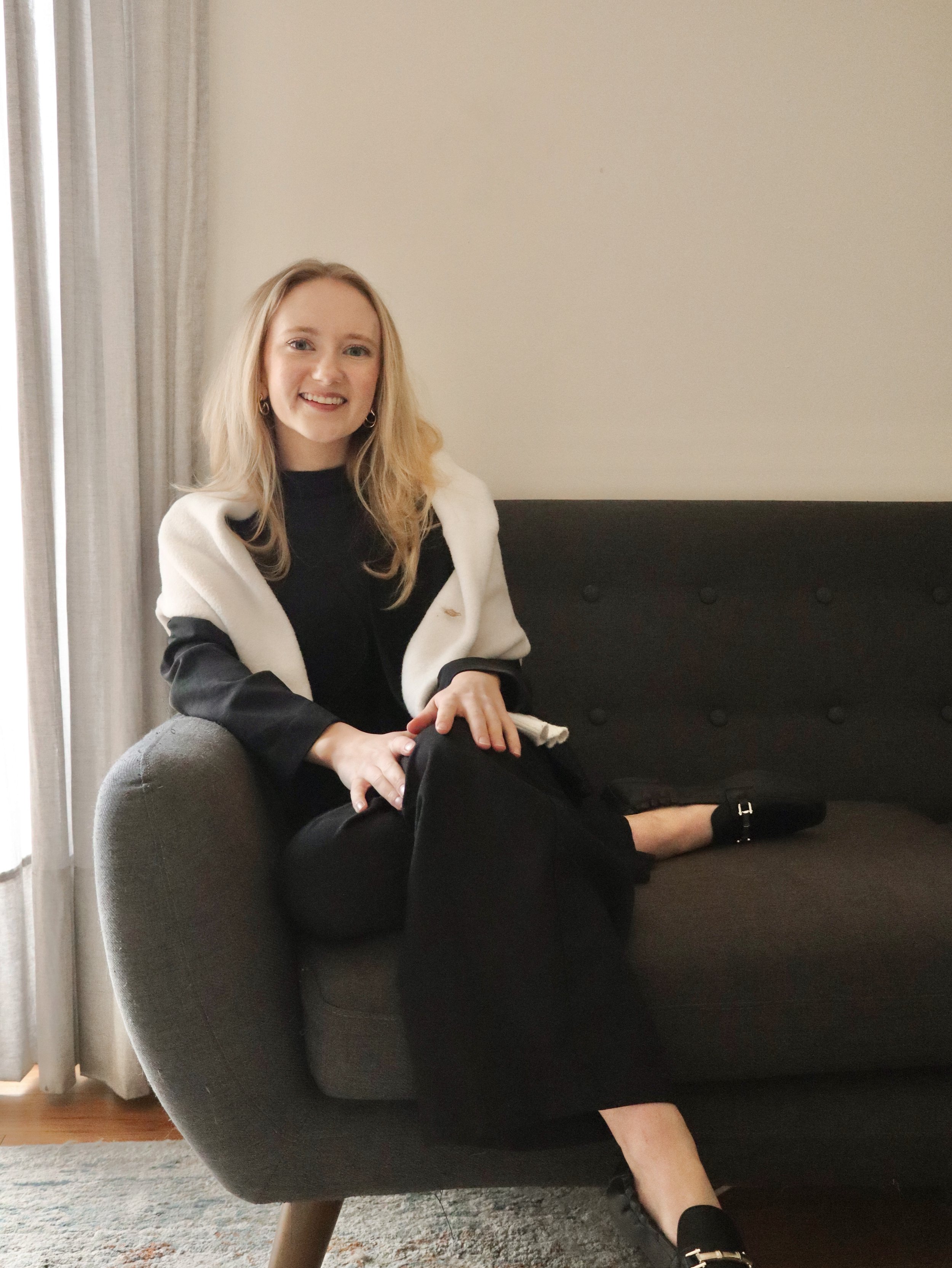 Smiling blonde woman in a black outfit and white cardigan sitting on a dark sofa beside a curtained window