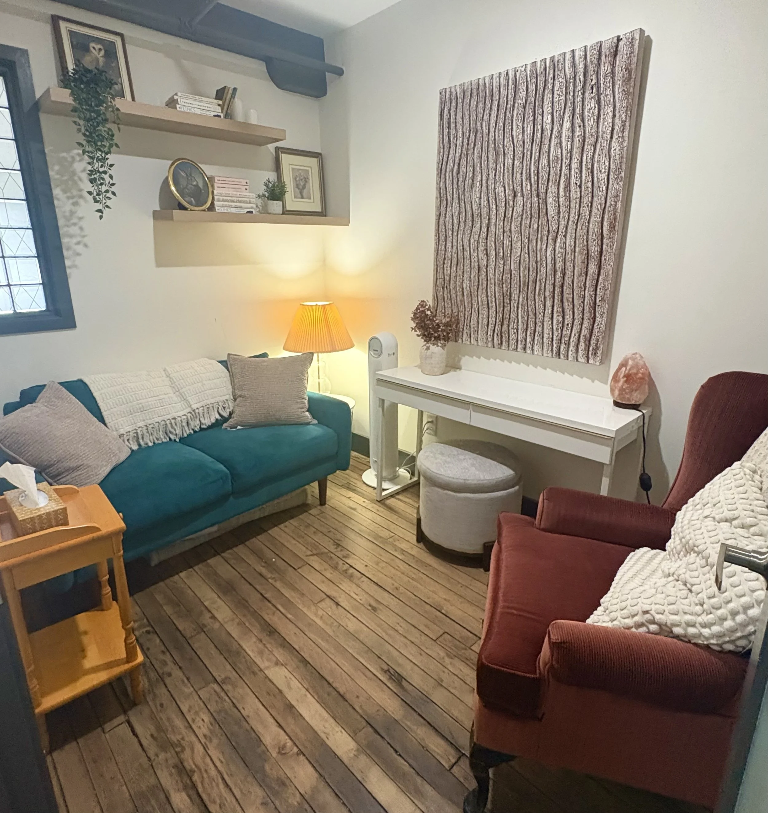 Cozy living room with a teal sofa, red armchair, white console table, illuminated lamp, framed artwork, shelves, and patterned wall paneling.