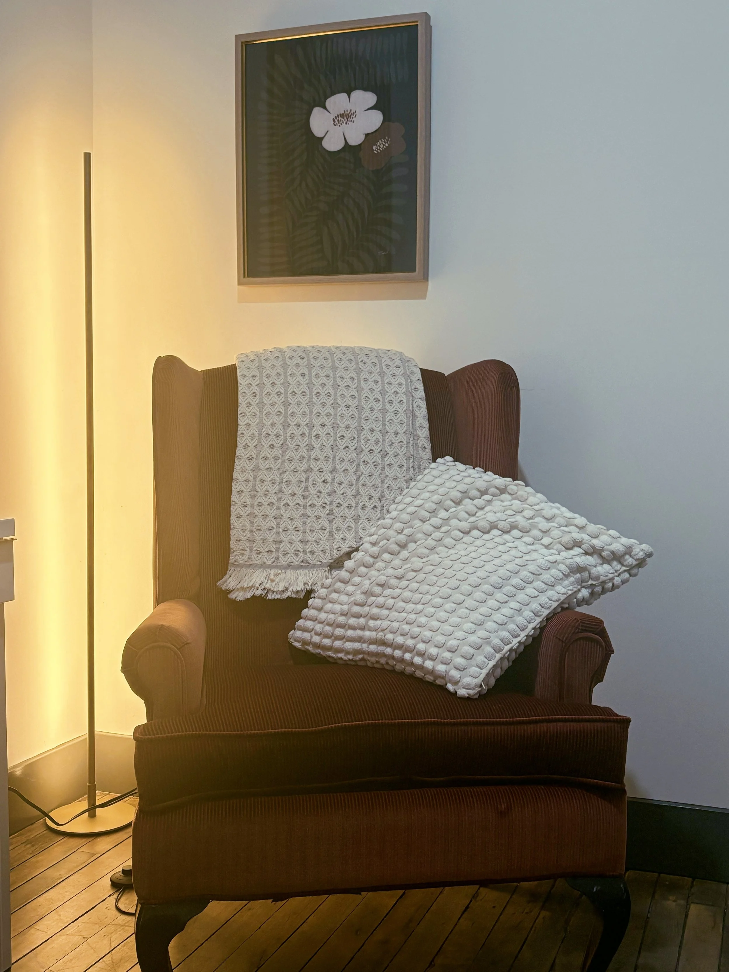Cozy brown armchair with textured white blankets beside a floor lamp and floral wall art