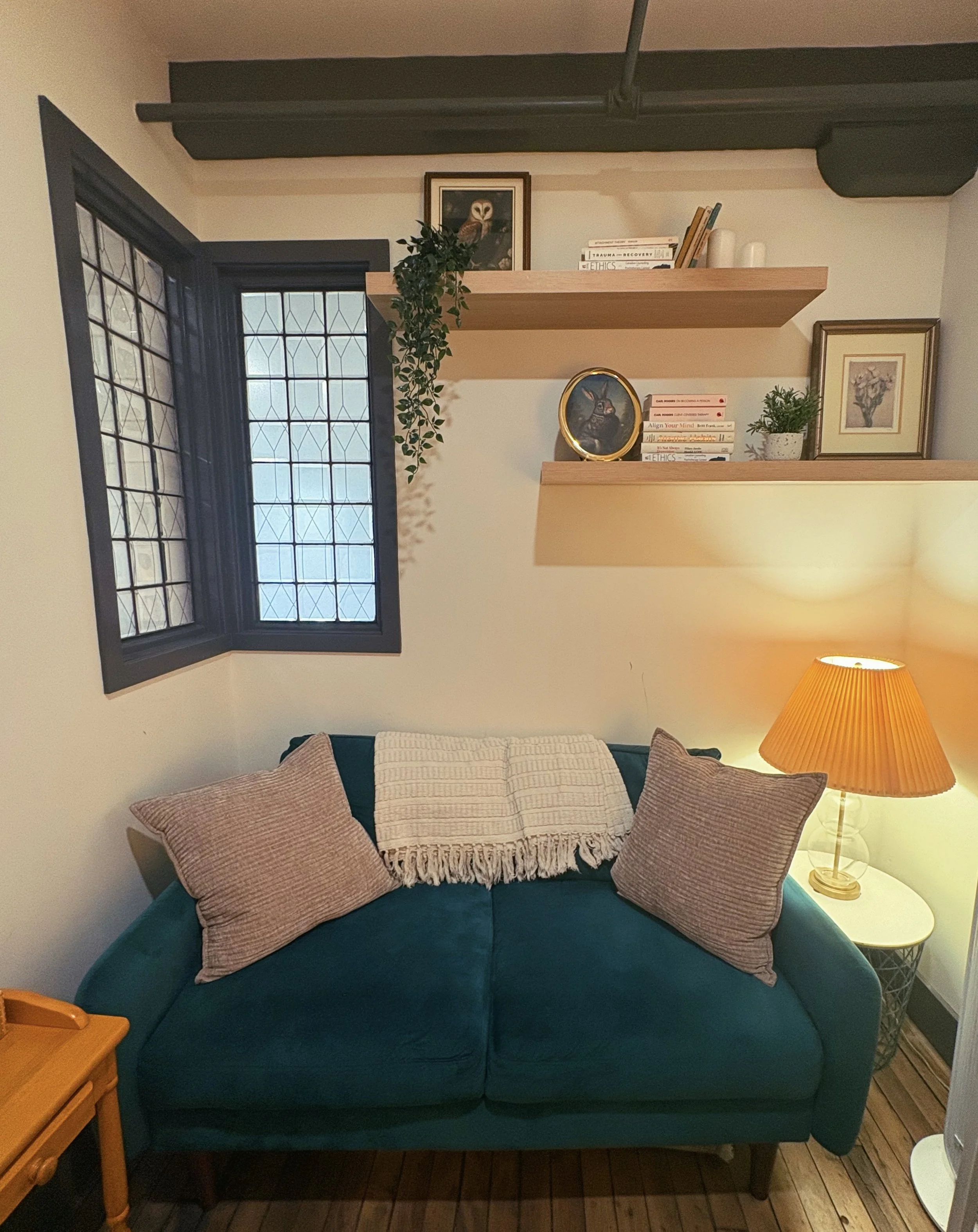 Cozy living room with a teal sofa, pink pillows, floating wooden shelves, books, framed art, plants, stained-glass window, and warm table lamp.