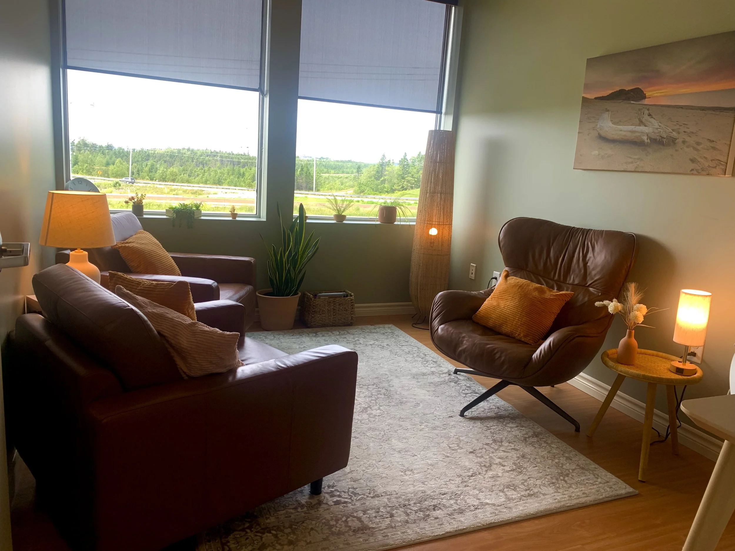 Cozy living room with leather armchair, sofa, side tables with lamps, large window with sunshine, green plants, and wall art of beach scene.