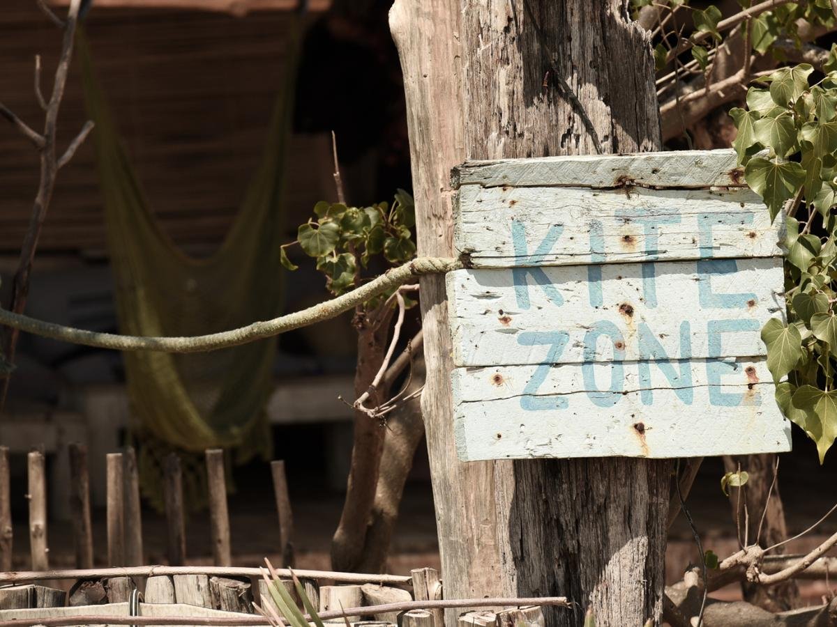 Cartello invecchiato con la scritta "Kite Zone" inchiodato al tronco di un albero, circondato da una rustica recinzione in legno e vegetazione. Sullo sfondo, un'amaca, a suggerire un'atmosfera rilassata.