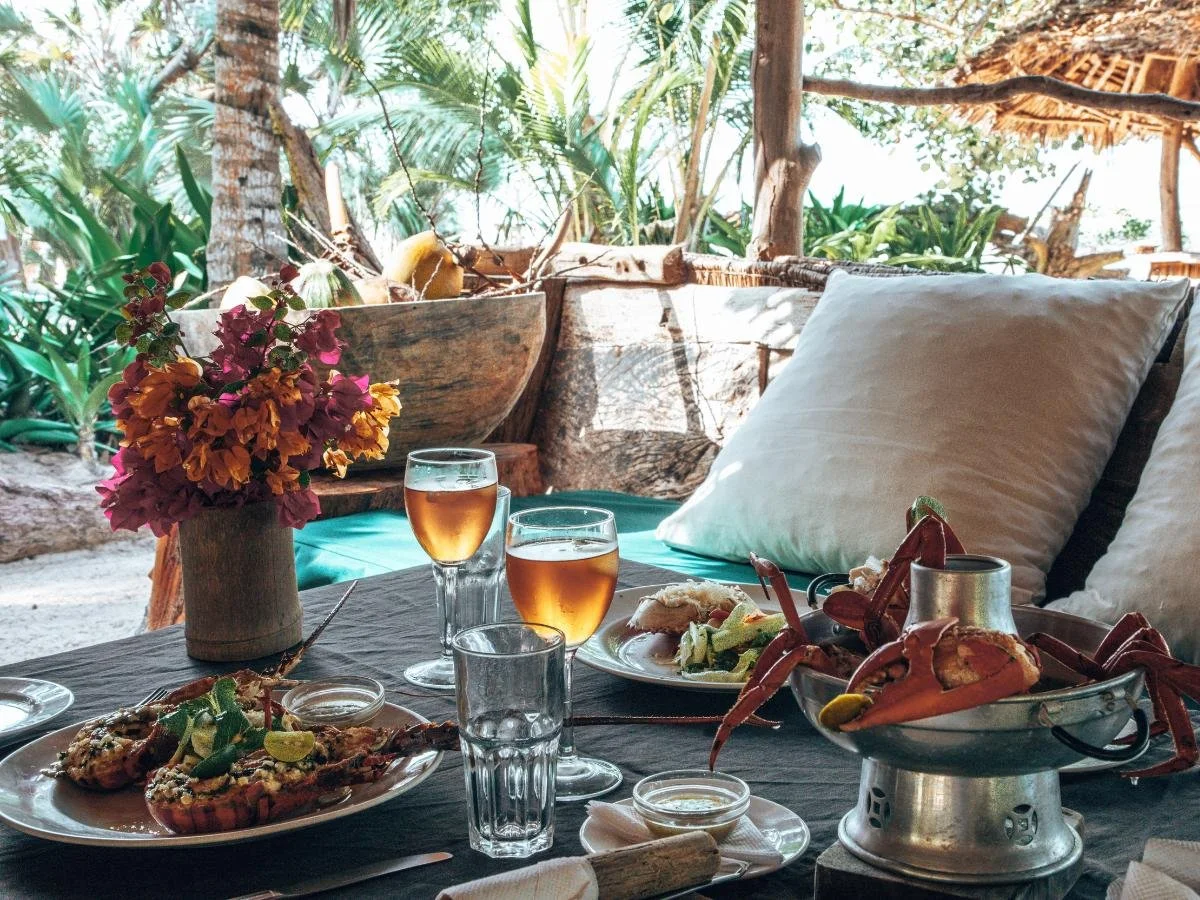 Scena di cena all'aperto con piatti di pesce, due bicchieri di bevande color ambra, vivaci fiori tropicali e una vegetazione lussureggiante sullo sfondo.