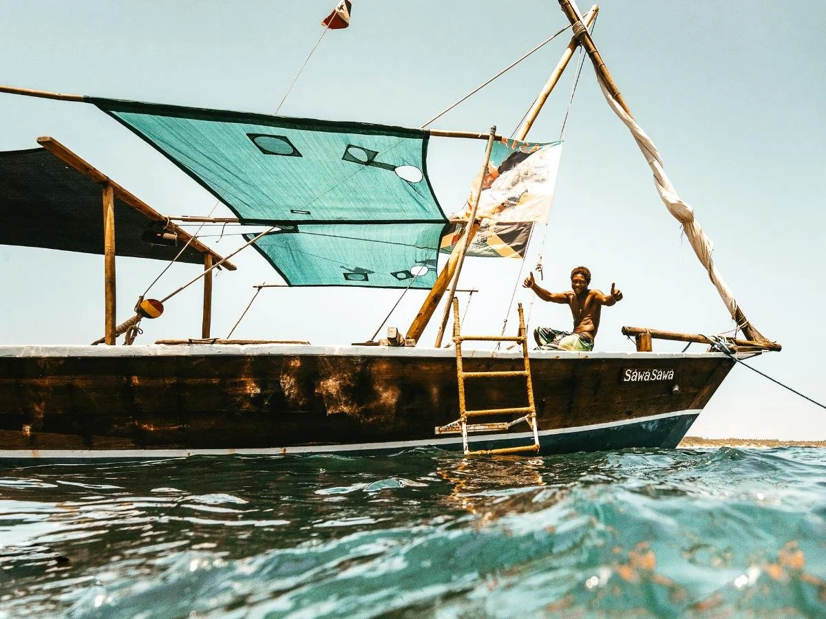 Un uomo posa gioiosamente su una piccola barca a vela di legno chiamata "Sawa Sawa" con le vele turchesi. La barca naviga su acque cristalline sotto un cielo luminoso.