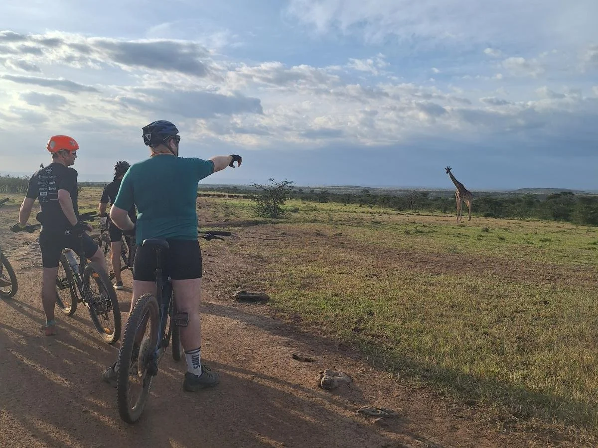 Tre ciclisti si fermano su un sentiero sterrato nella savana; uno di loro indica una giraffa che cammina a poca distanza nella pianura erbosa. Il cielo è parzialmente nuvoloso con la luce morbida del tardo pomeriggio.