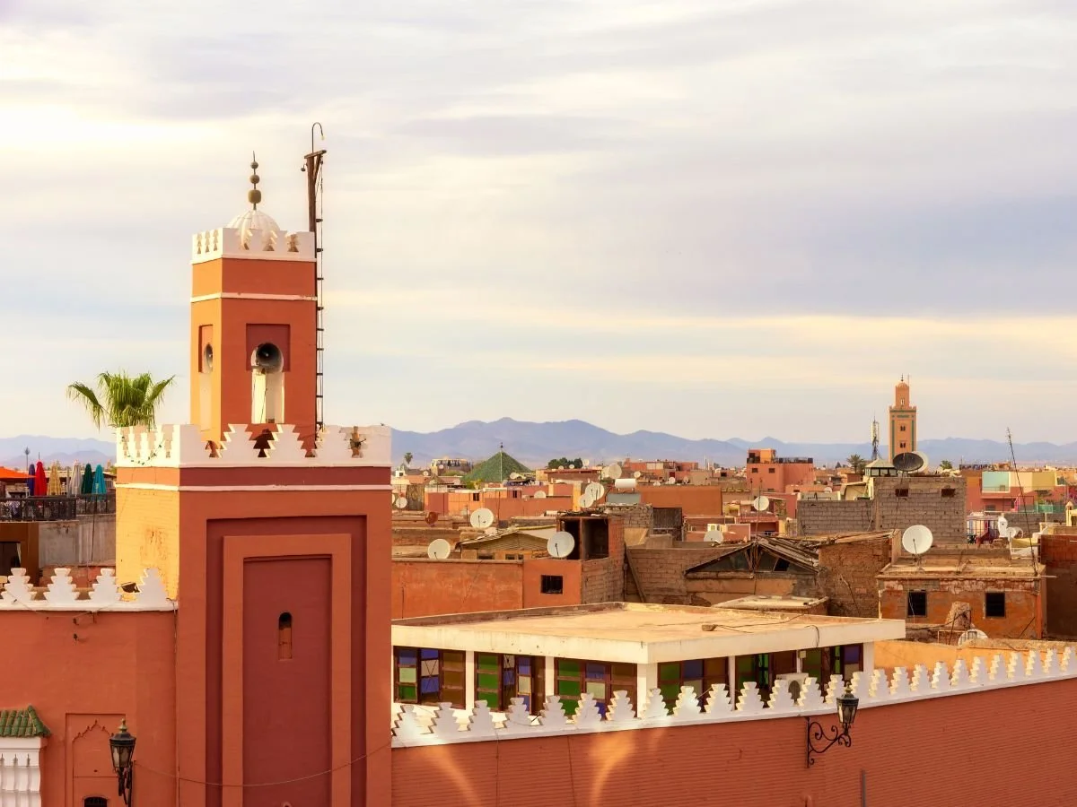 Vista panoramica sui tetti in terracotta e i minareti di Marrakech al tramonto con le montagne dell'Atlante all'orizzonte.