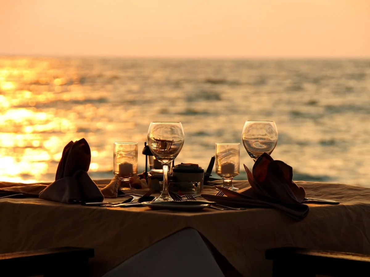 Cena romantica in riva al mare al tramonto. Tavolo con calici da vino, candele e tovaglioli, con vista sull'oceano scintillante. Atmosfera calda e tranquilla.