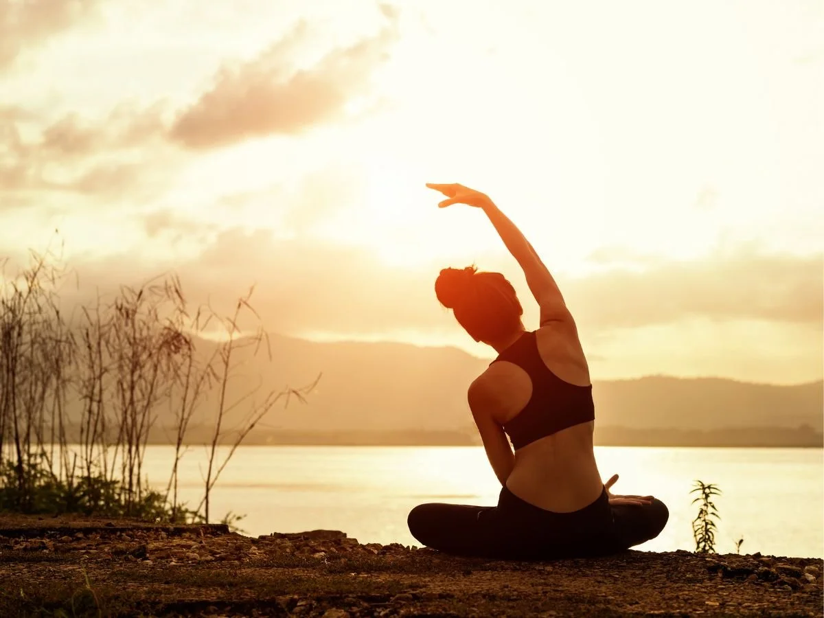 Una donna seduta in posizione yoga allunga il braccio sopra la testa all'alba, in riva a un lago tranquillo. La silhouette si staglia contro un cielo luminoso, evocando calma e serenità.