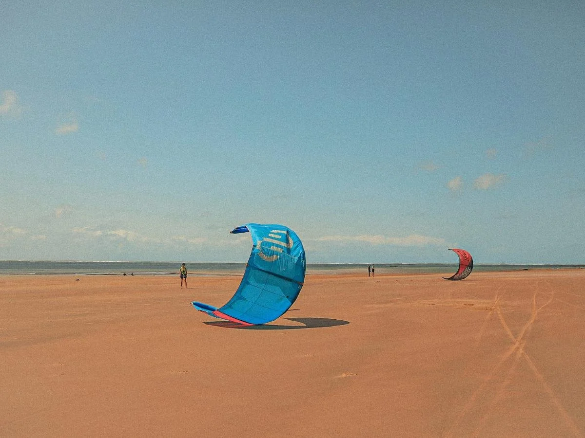 Una vasta spiaggia sabbiosa con due colorati aquiloni da kitesurf a terra. Il mare è calmo sotto un cielo azzurro e limpido, con qualche persona in lontananza.