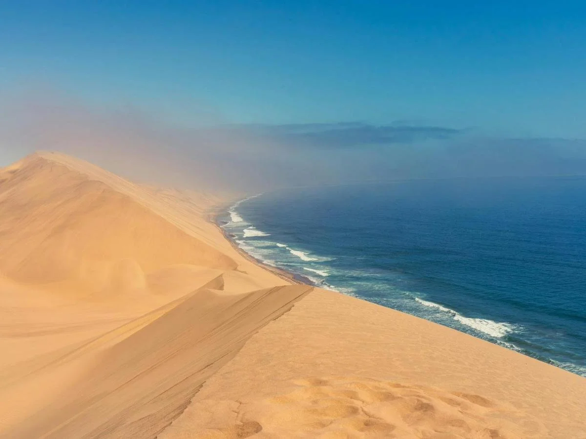 Le imponenti dune di sabbia dorata di Sandwich Harbour che si tuffano direttamente nelle acque dell'Oceano Atlantico sotto un cielo azzurro.
