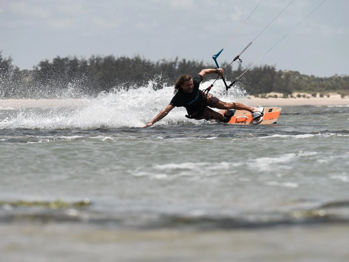 Un kitesurfer scivola abilmente sull'acqua, appoggiandosi all'indietro e toccando la superficie con una mano. Sullo sfondo, alberi e una spiaggia sabbiosa.