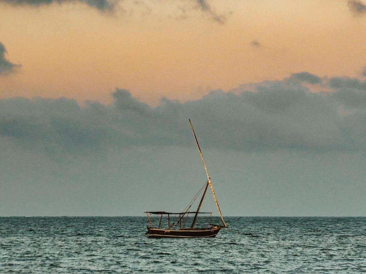 Una piccola barca a vela è adagiata sulle calme acque dell'oceano sotto un cielo color pesca pastello e grigio, evocando un'atmosfera serena e tranquilla al tramonto.