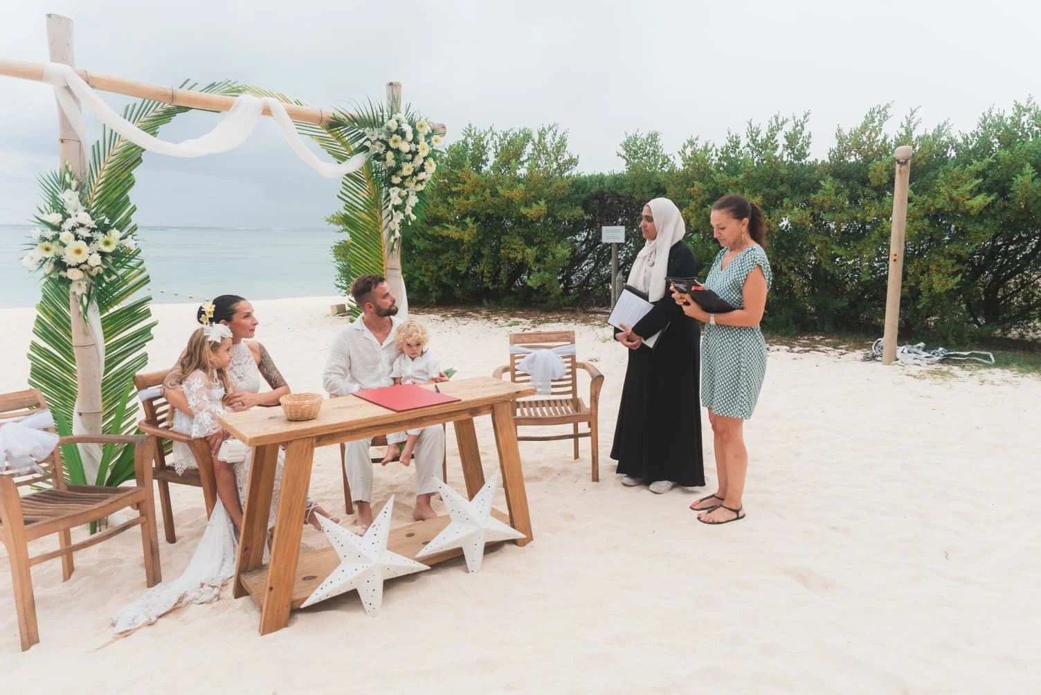 Una coppia con due bambini è seduta a un tavolo di legno su una spiaggia, con un arco decorato e l'oceano alle spalle. Una donna e il celebrante sono in piedi nelle vicinanze, creando una scena nuziale serena.